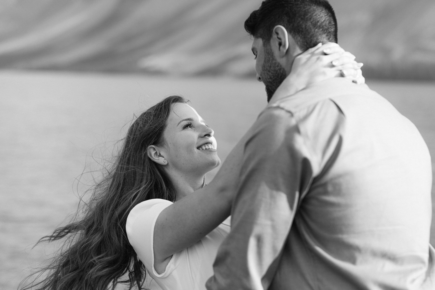 Black and white image of a woman smiling joyously as she looks up at a man while embracing him, with a blurred background of a lake and hills.