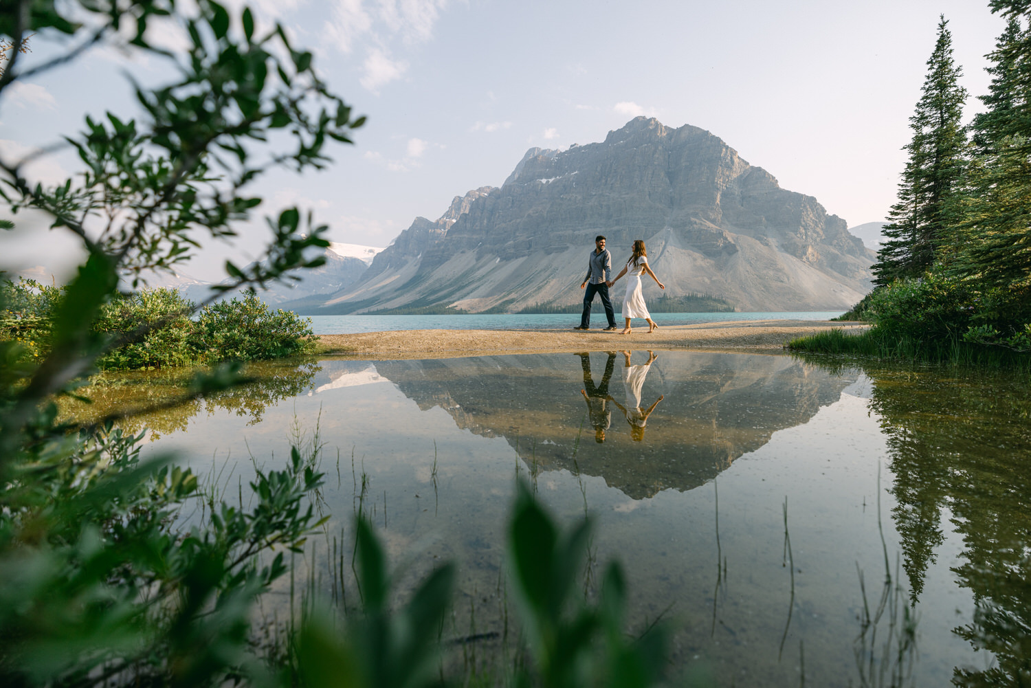 Two people holding hands and walking by a tranquil mountain lake with their reflection on the water.