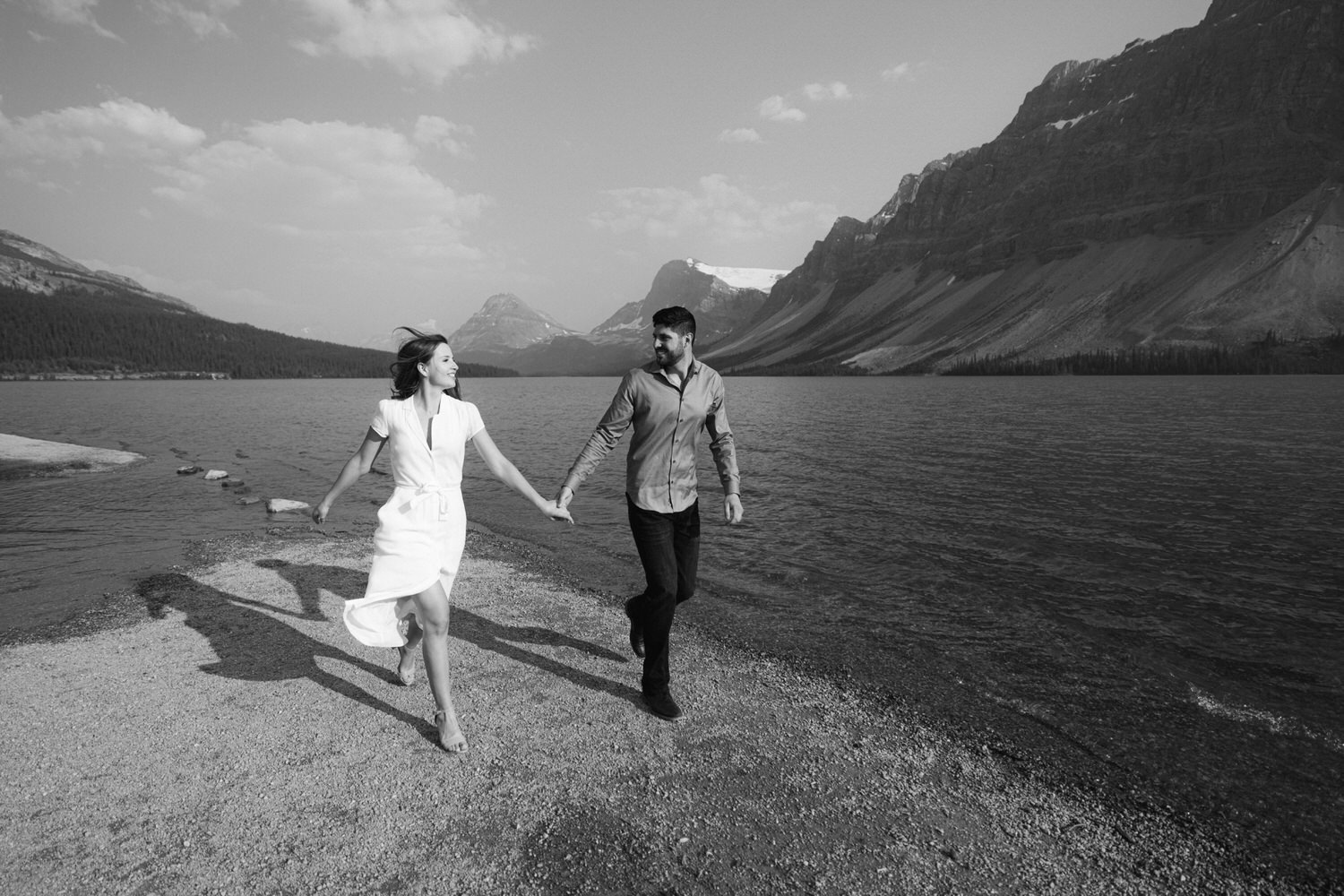 A black and white photo of a couple holding hands and running joyfully along a lakeshore with mountains in the background.