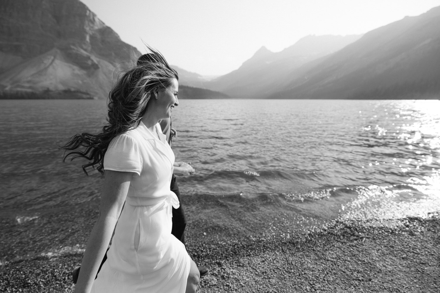 A woman in a white dress standing by a mountain lake with her hair blowing in the wind, in a black and white setting.