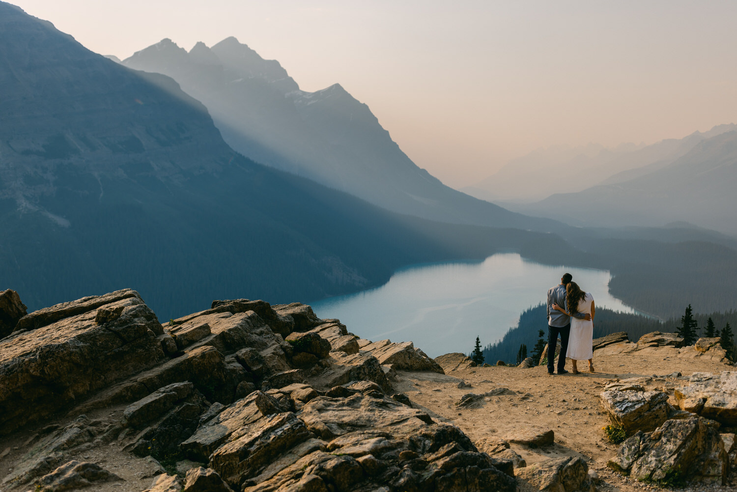 A couple embracing while looking at a mountainous landscape with a lake below, in hazy sunlight.