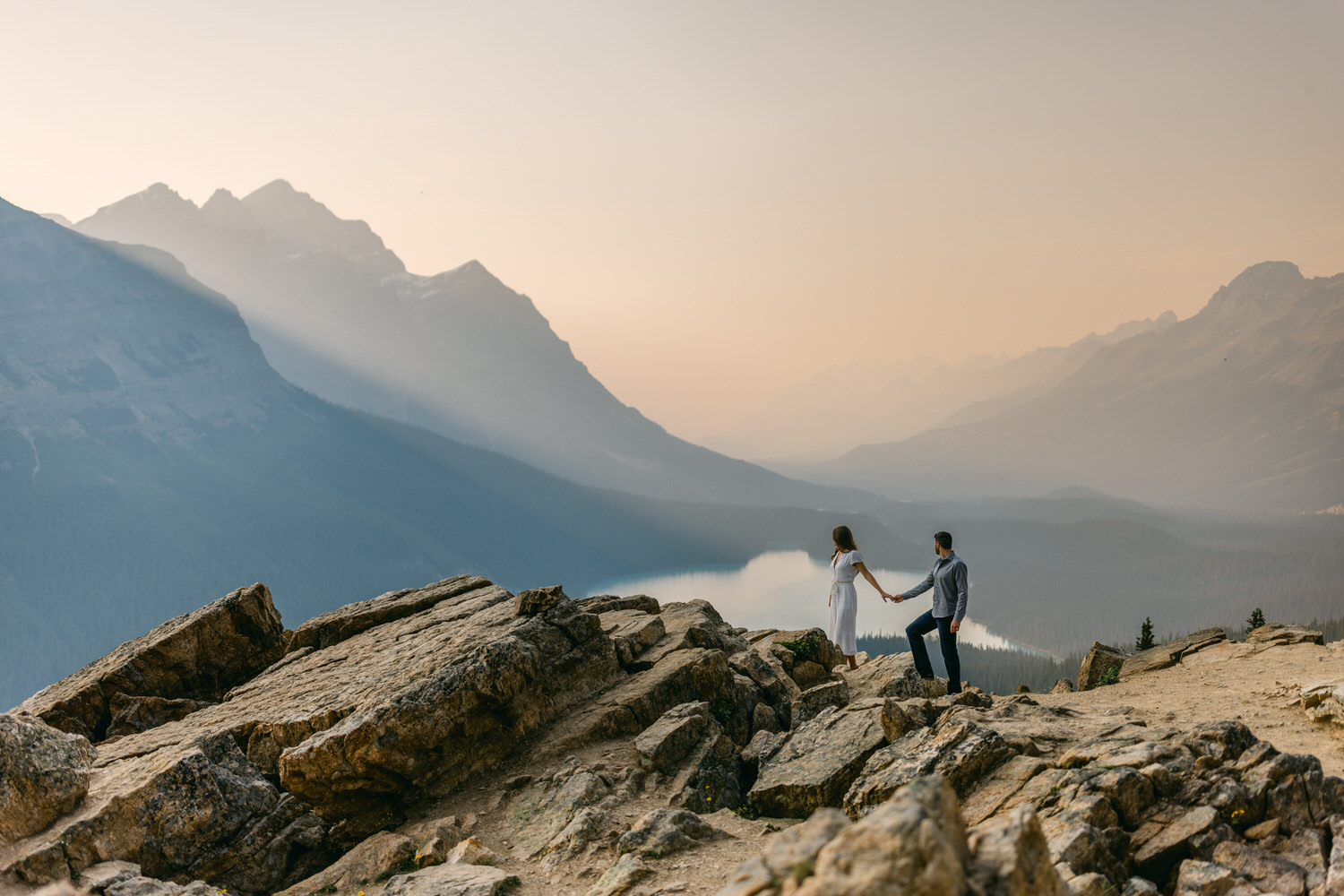 A couple holding hands on a rocky overlook with a serene mountain range and lake in the background at dusk.