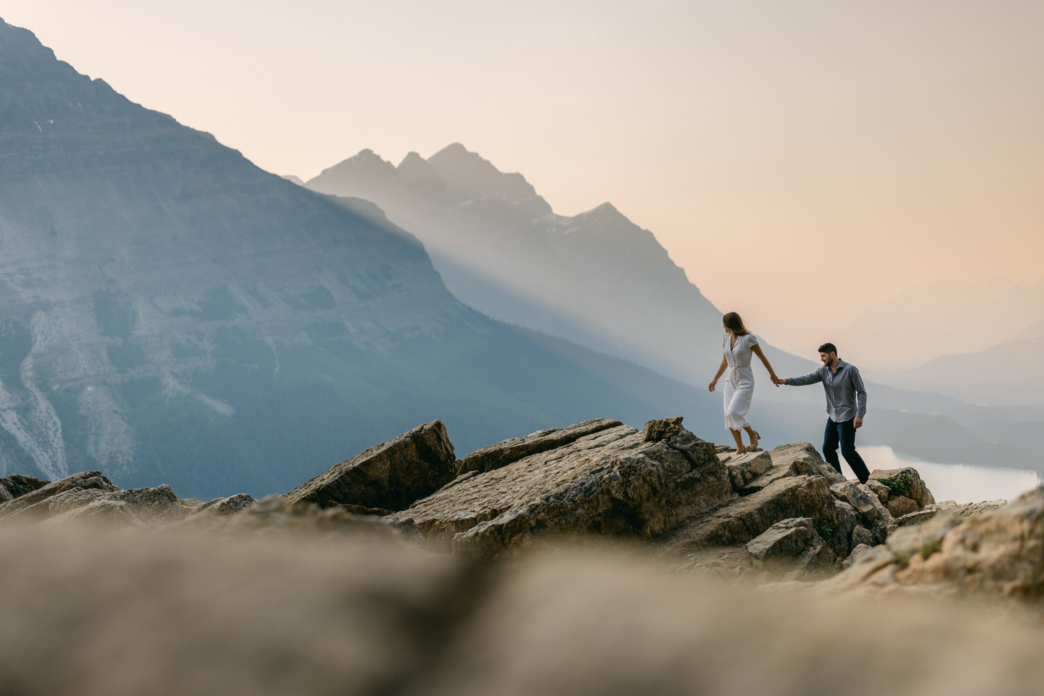 A man and woman in casual attire walking hand in hand over a rocky outcrop, with layered mountain silhouettes in the hazy background.