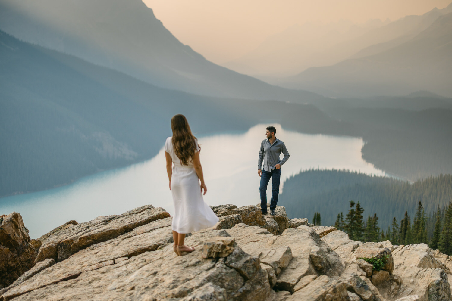 A woman in a white dress walking towards a man standing on a rocky overlook with a scenic mountain lake in the background at sunset