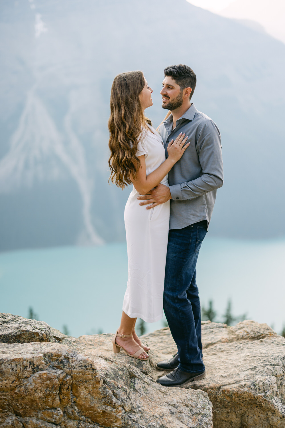 A couple holding each other on a rocky outcrop with a turquoise lake and mountains in the background.