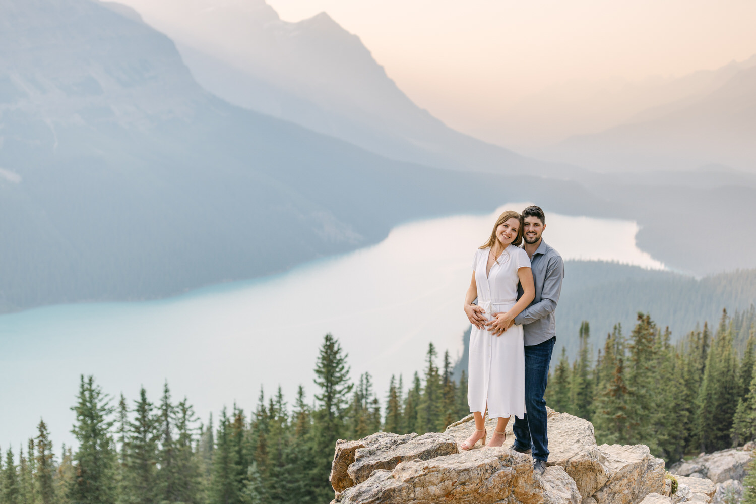 A couple standing close and smiling on a mountain overlooking a turquoise lake with pine trees and hazy mountains in the background