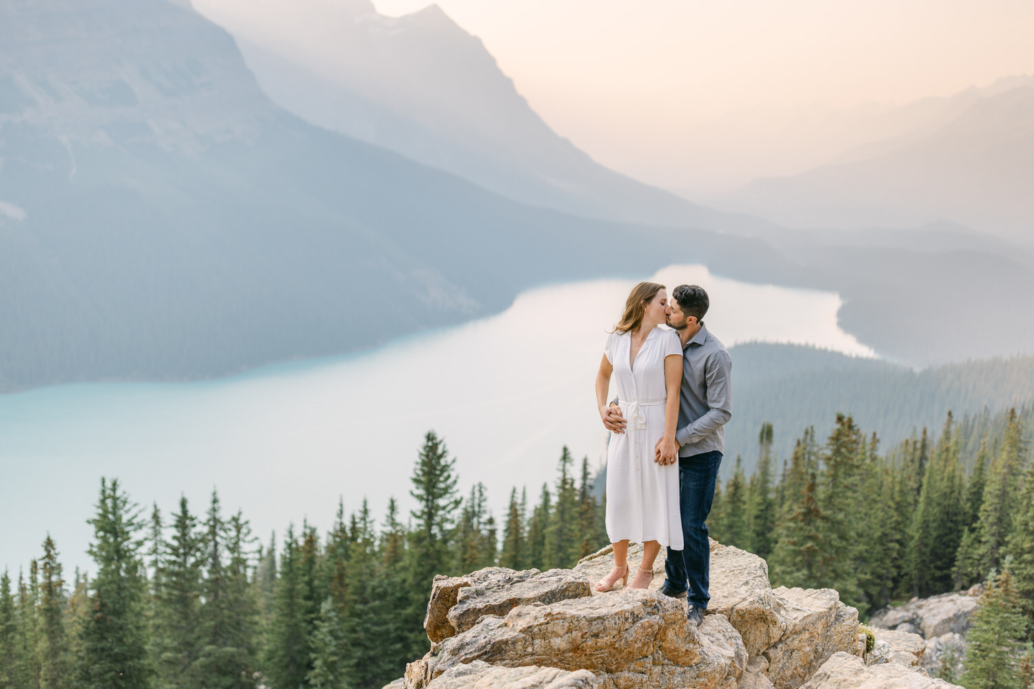 A couple sharing a kiss on a rocky overlook with a tranquil mountain lake and forest in the background