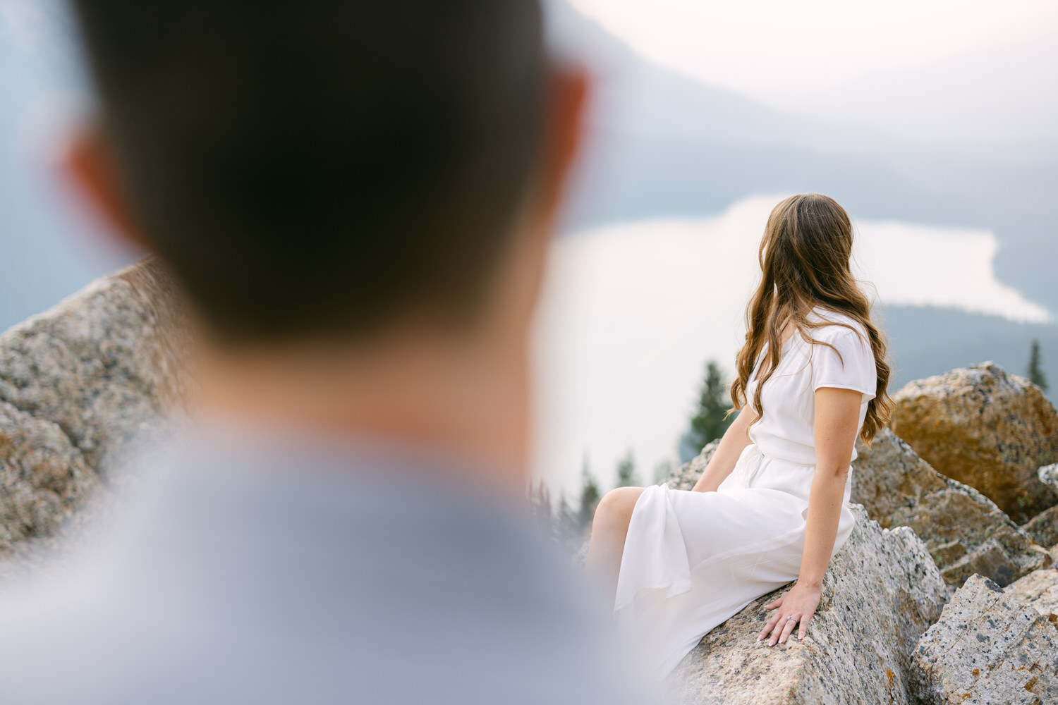 A person in a white dress sitting on a rocky overlook with a blurry figure in the foreground and a lake in the distance.