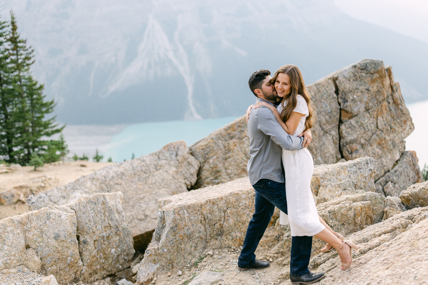 A couple hugging on a rocky outcrop with a turquoise lake and pine trees in the blurry background