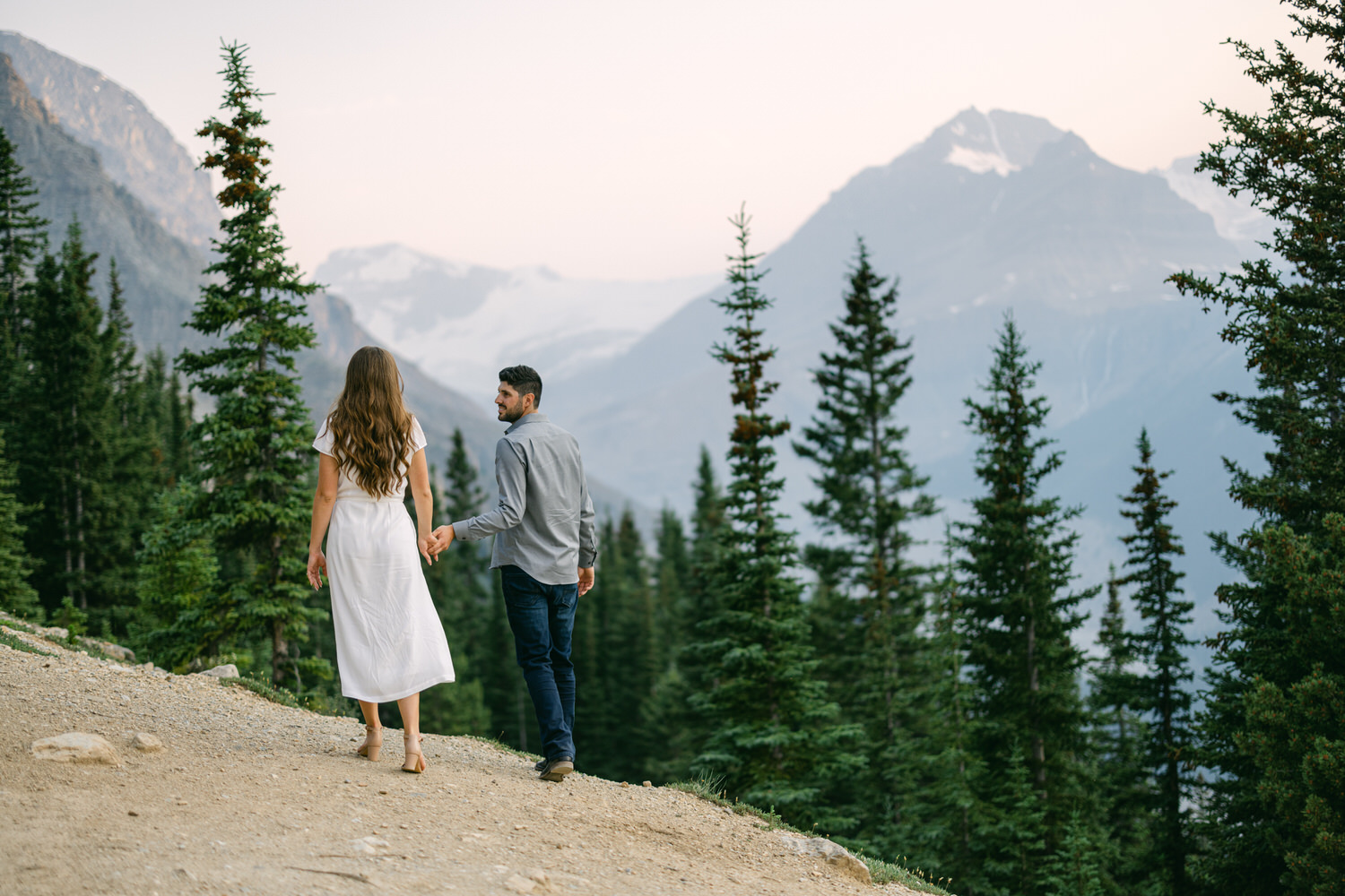 A couple holding hands while walking on a mountain trail surrounded by evergreen trees with a mountainous backdrop.