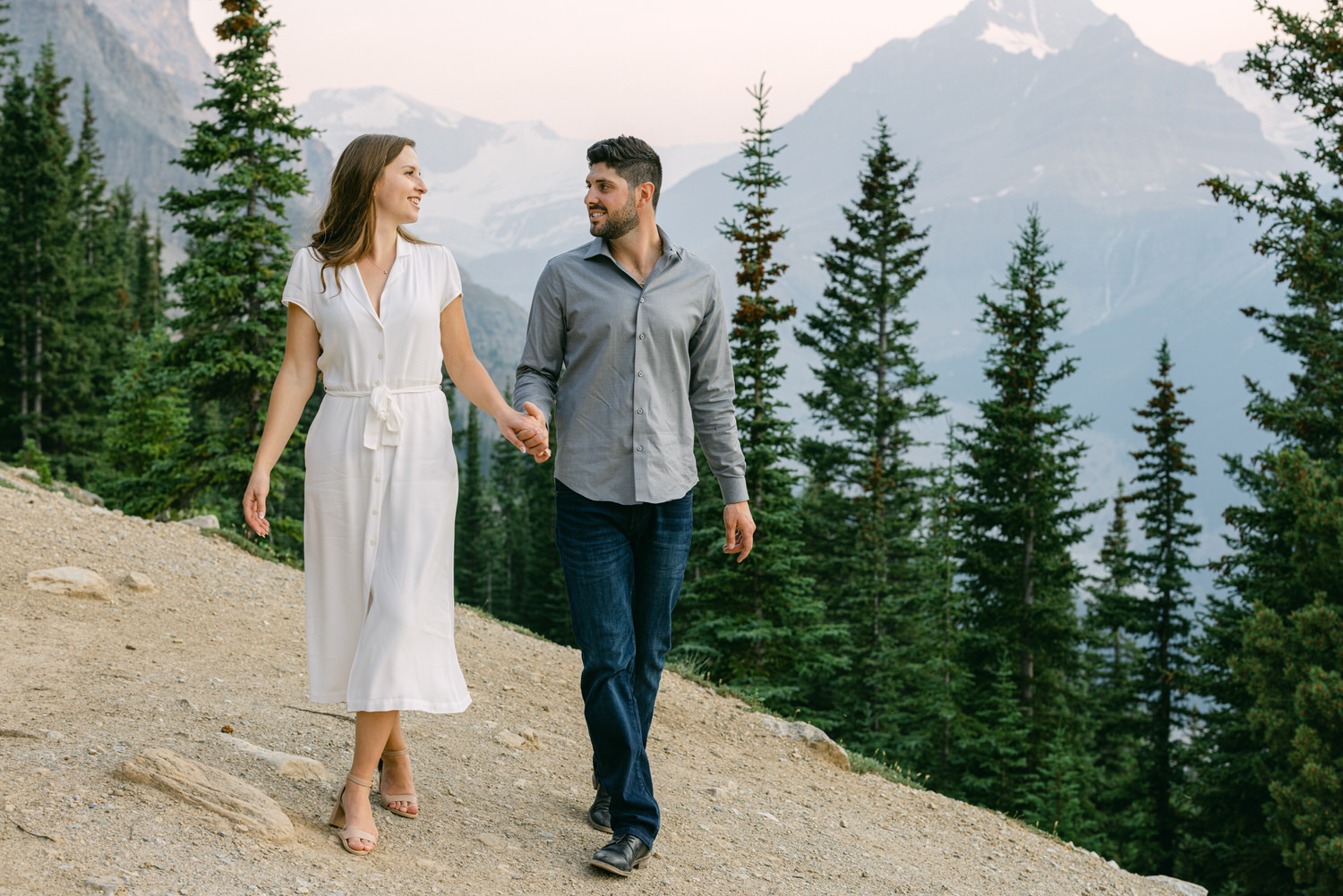 A couple holding hands and walking along a mountain trail with evergreen trees and mountains in the background.