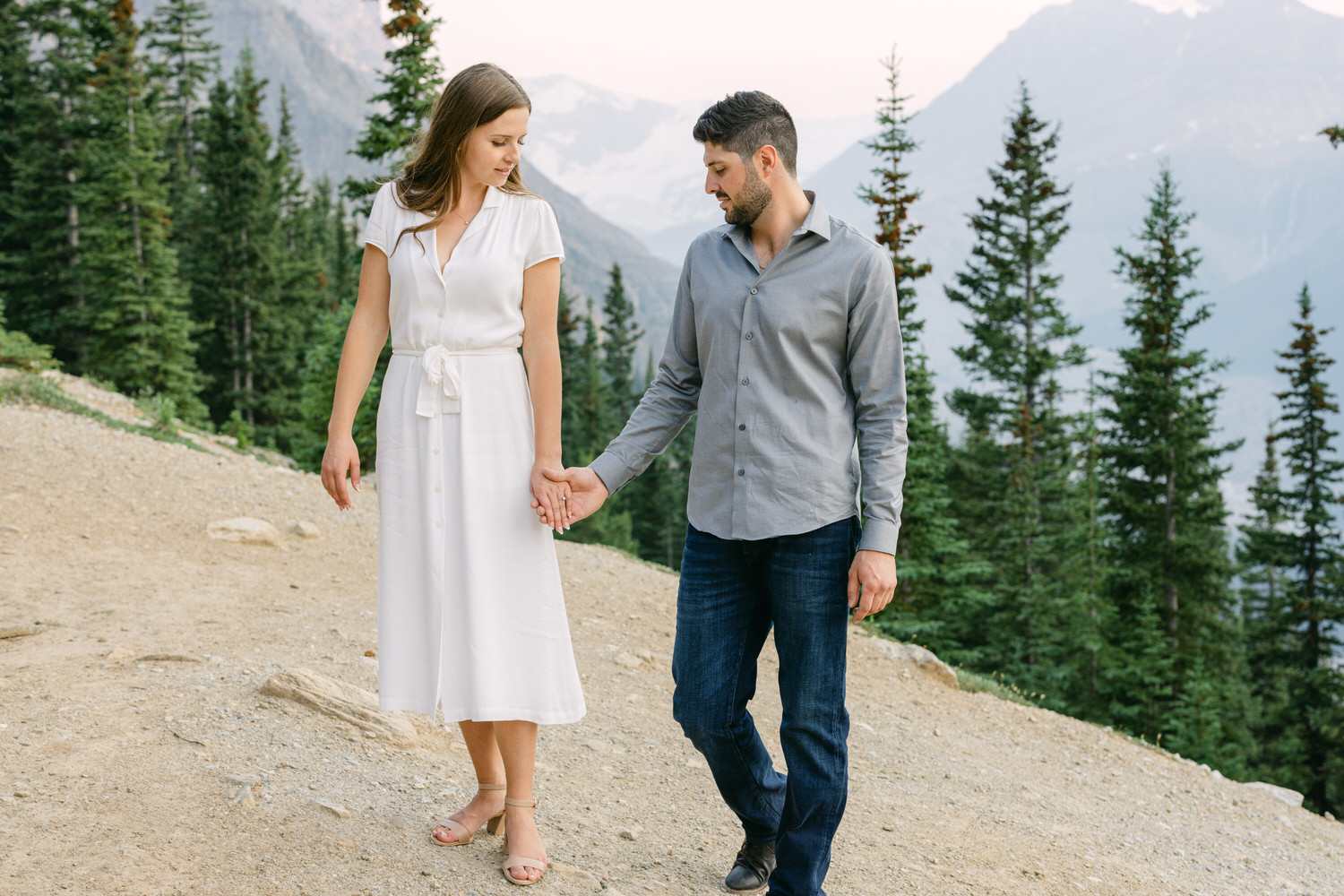 A man and a woman holding hands and walking together on a mountain trail surrounded by pine trees.