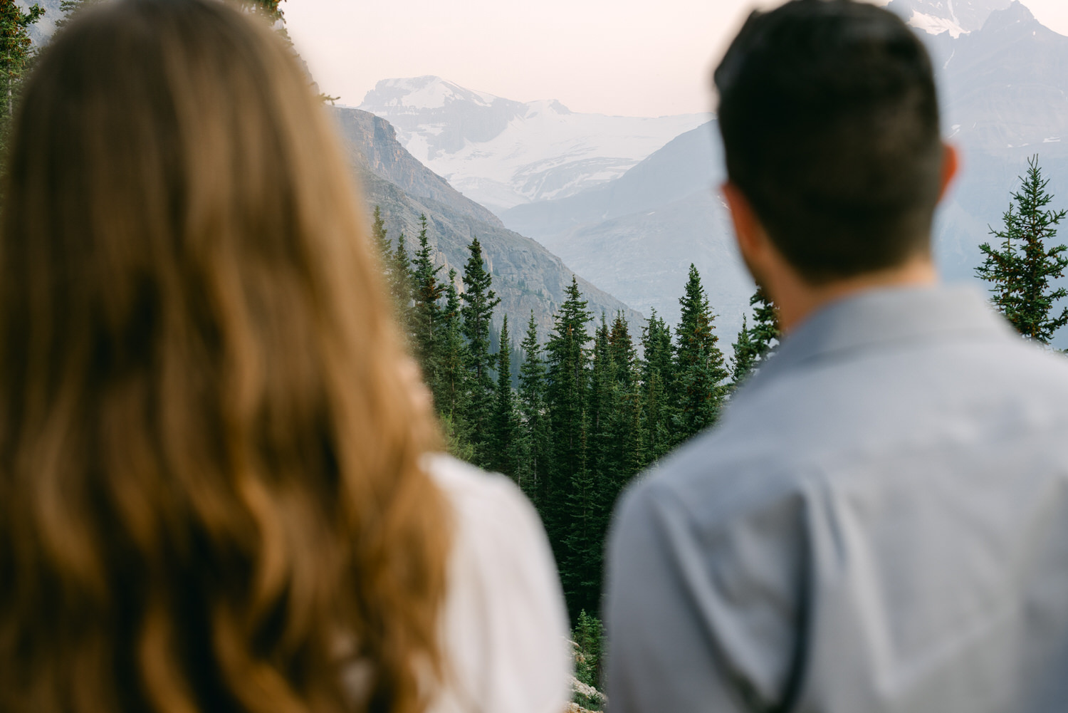 Two individuals from behind looking at a mountainous landscape with pine trees.