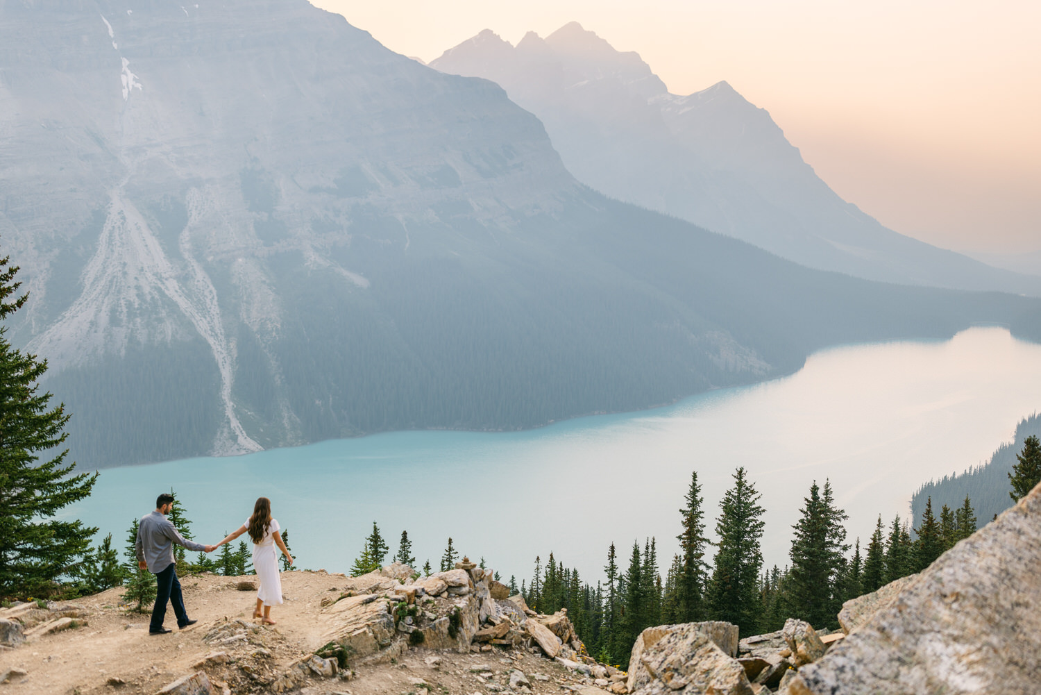 A couple holding hands while standing on a rocky overlook with a panoramic view of a turquoise mountain lake and pine trees, with mountains in the hazy background during sunset.