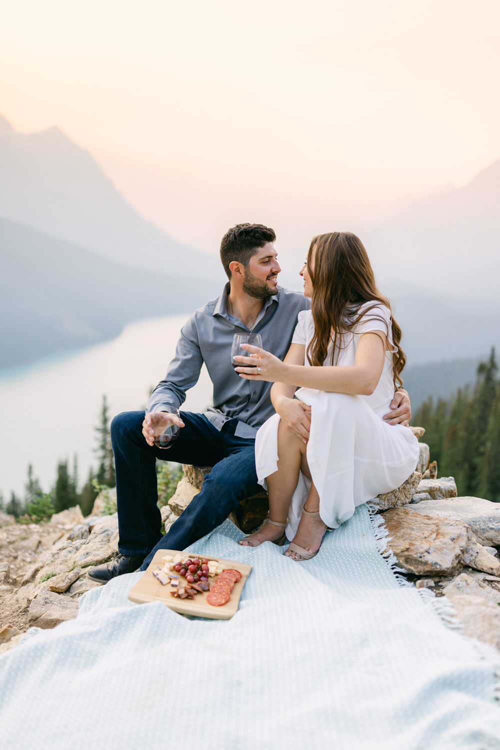 A couple enjoying a romantic picnic with a cheese and fruit platter on a blanket, overlooking a mountainous landscape.