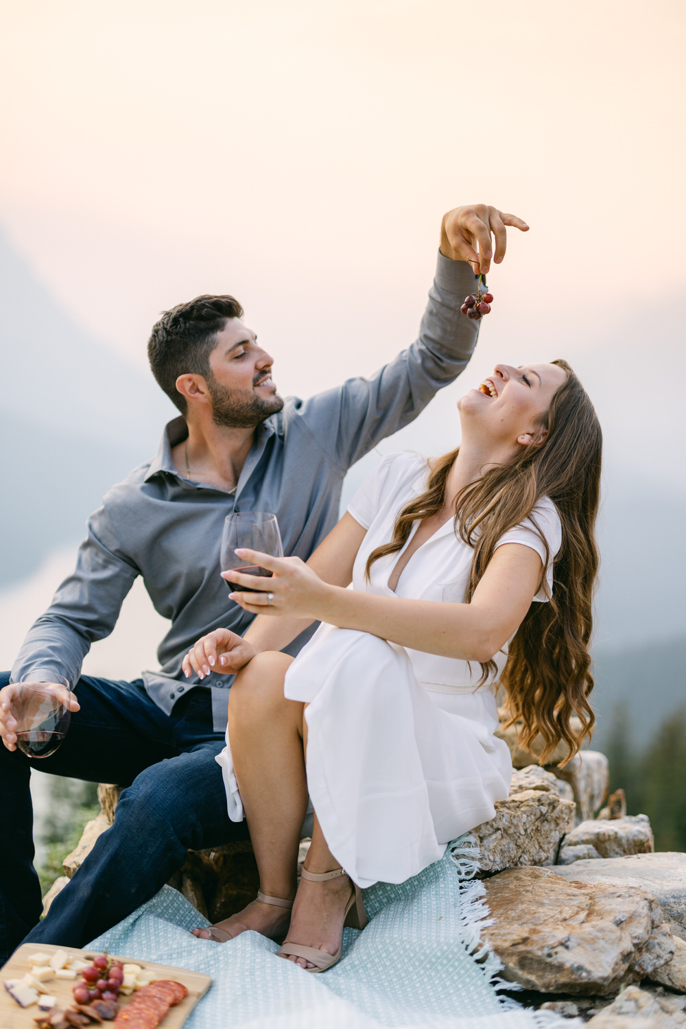 A couple enjoying a playful moment sharing grapes during a mountain picnic with wine and cheese platter.