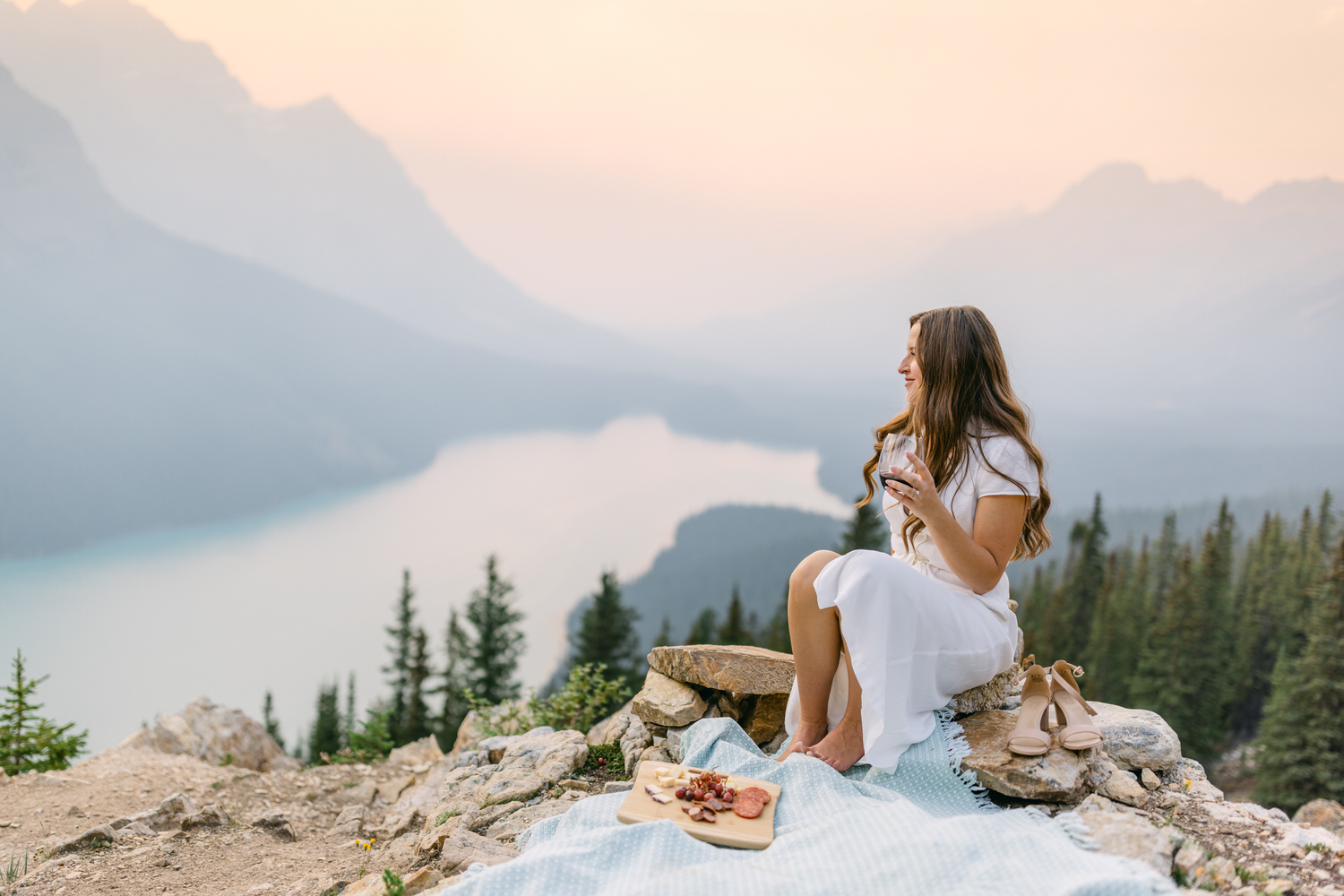 A woman in a white dress sitting on a rock, enjoying a picnic with a scenic mountain lake view in the background.