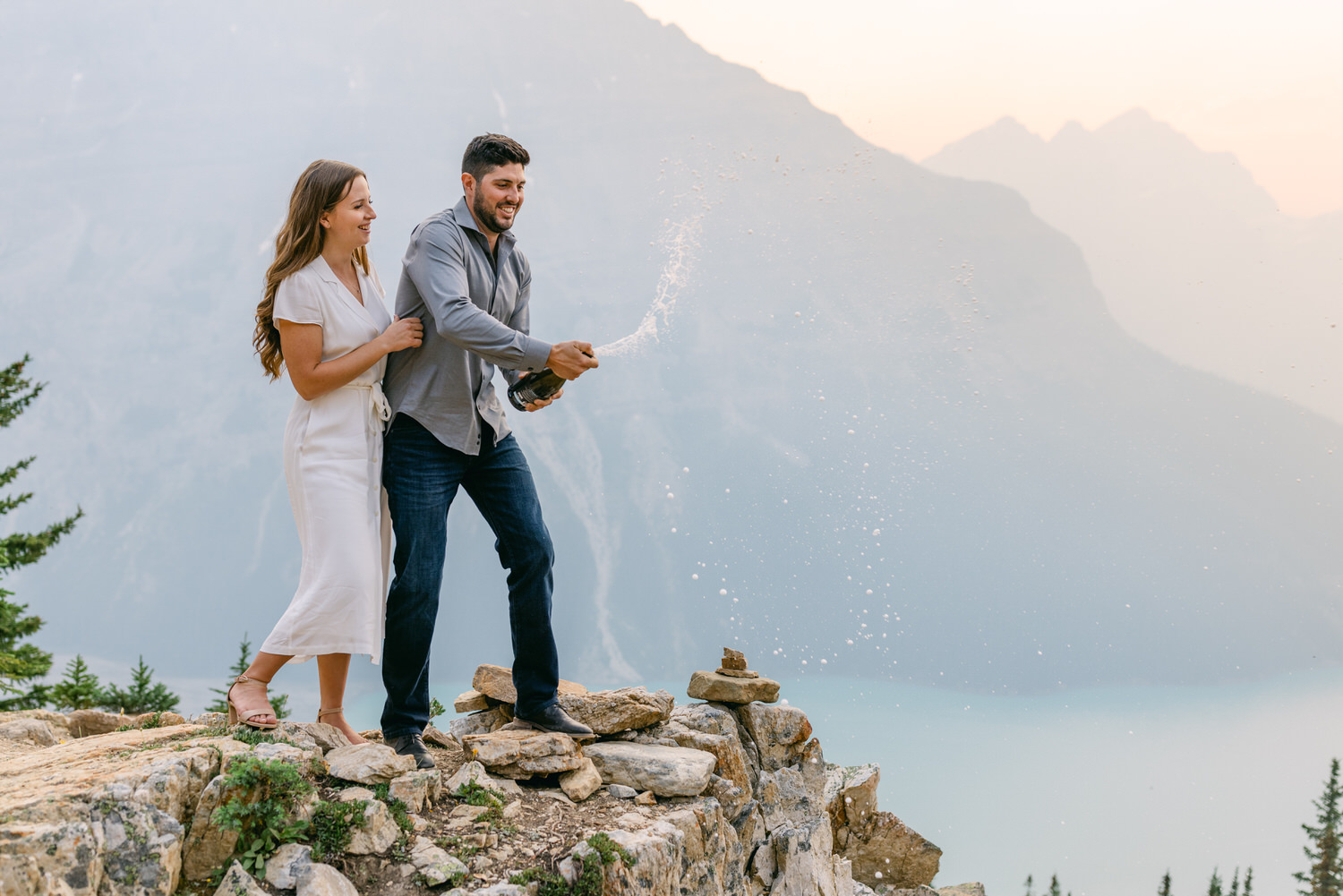 A man and woman popping a bottle of champagne on a mountain overlooking a scenic view