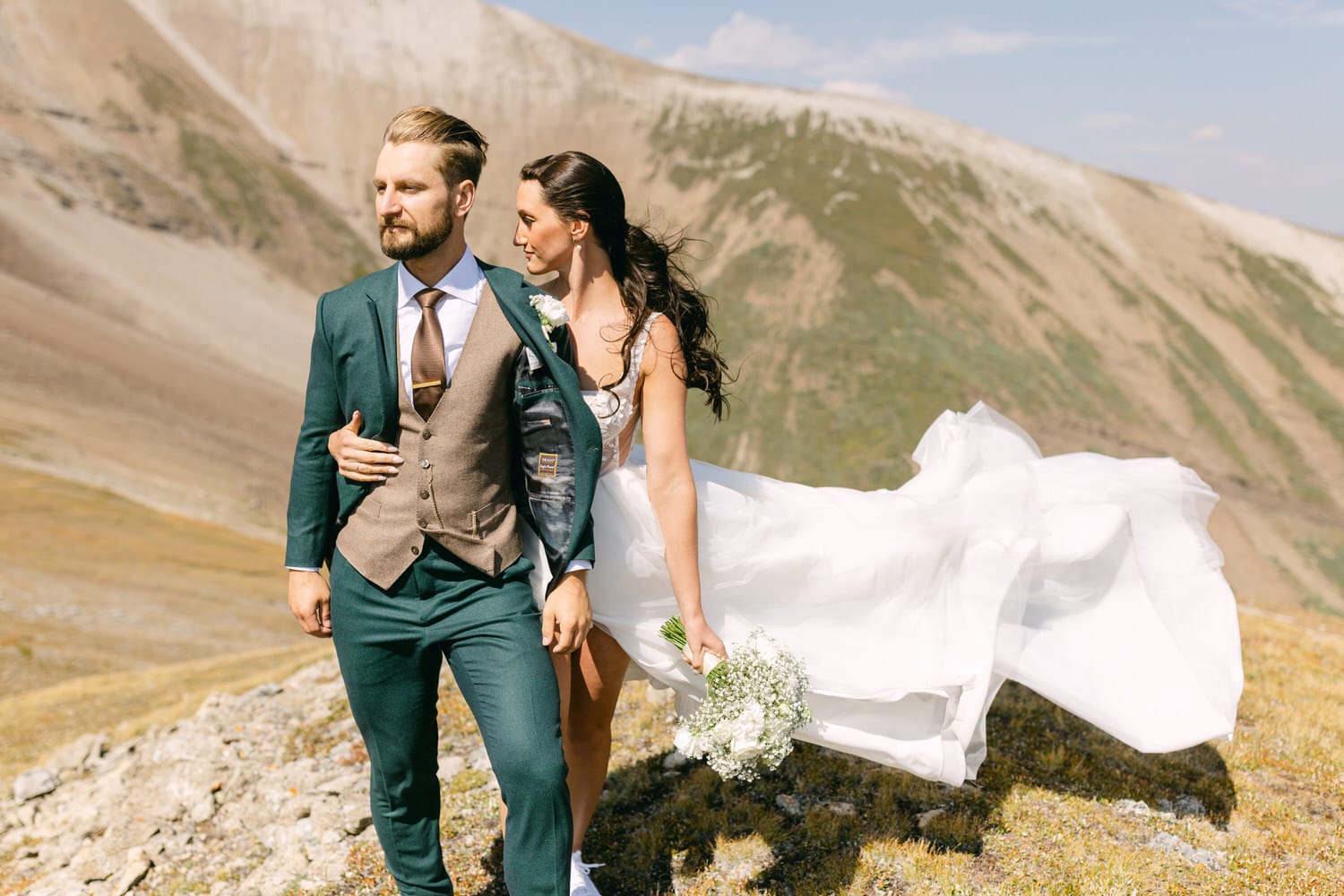 A bride and groom in elegant attire standing against a backdrop of mountains, with the bride's flowing dress caught in a gentle breeze.