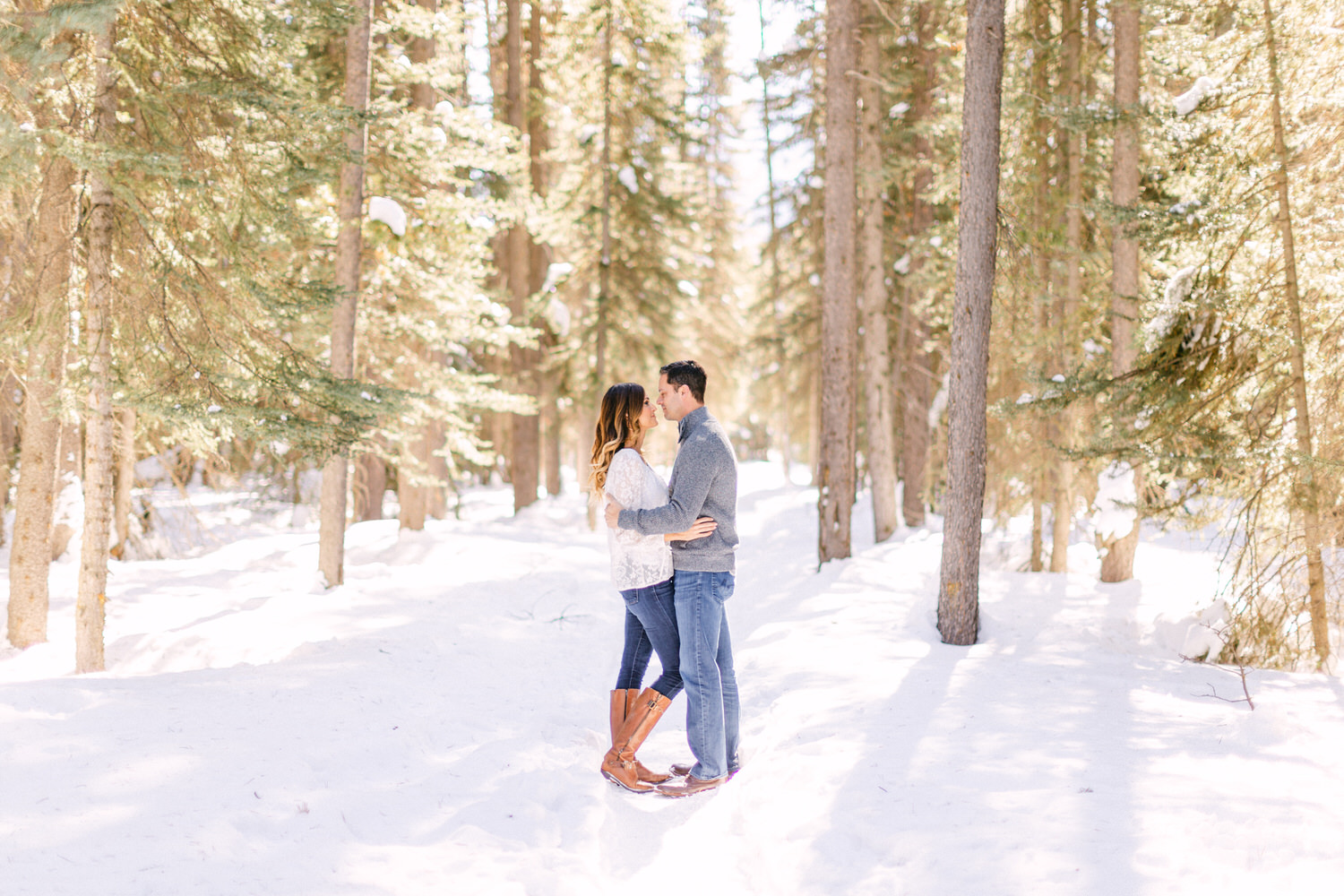 A couple standing close together surrounded by snow-covered trees in a sunlit forest.