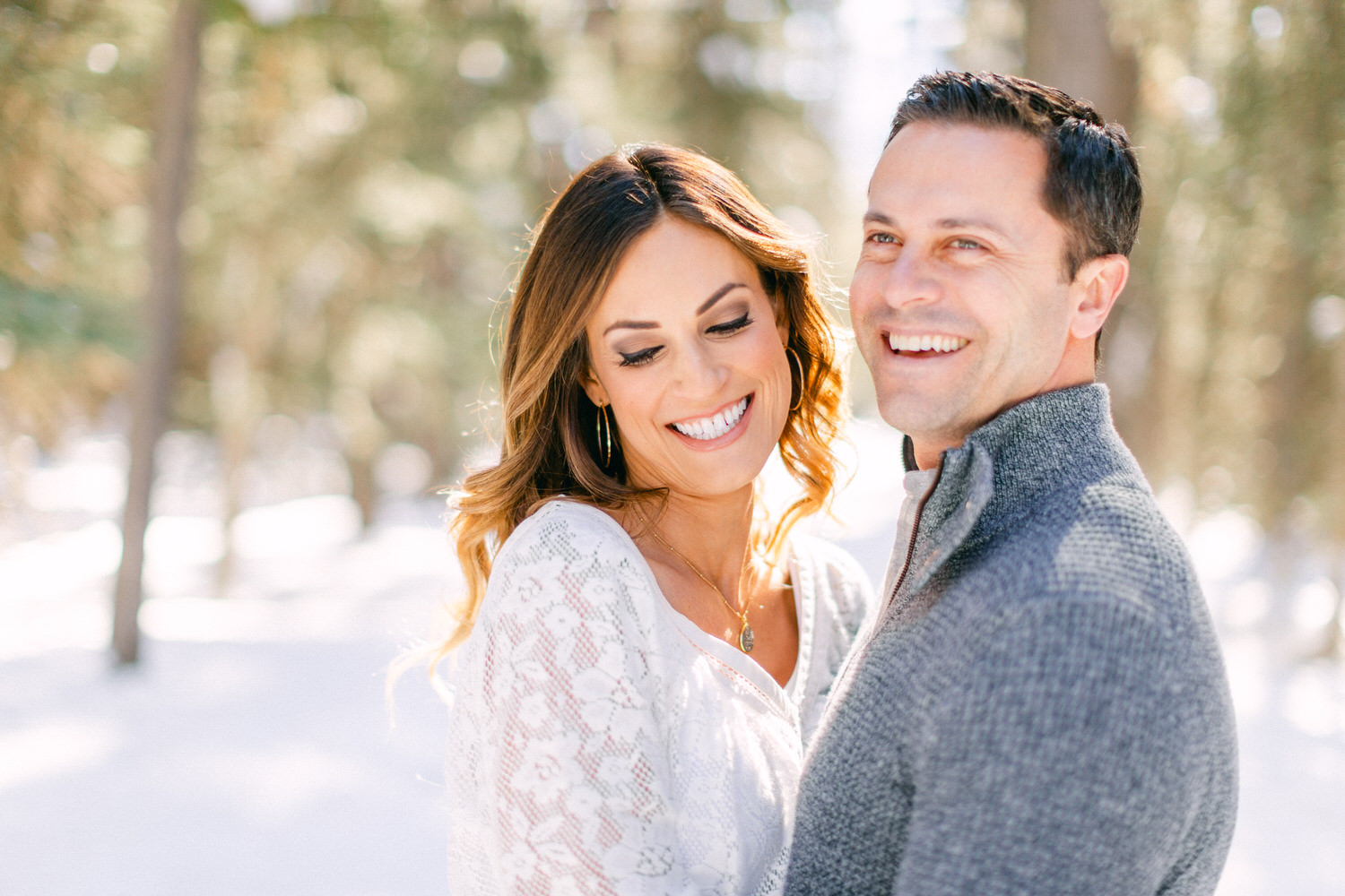 A man and woman smiling and embracing each other in a snowy landscape, with sun rays filtering through trees in the background