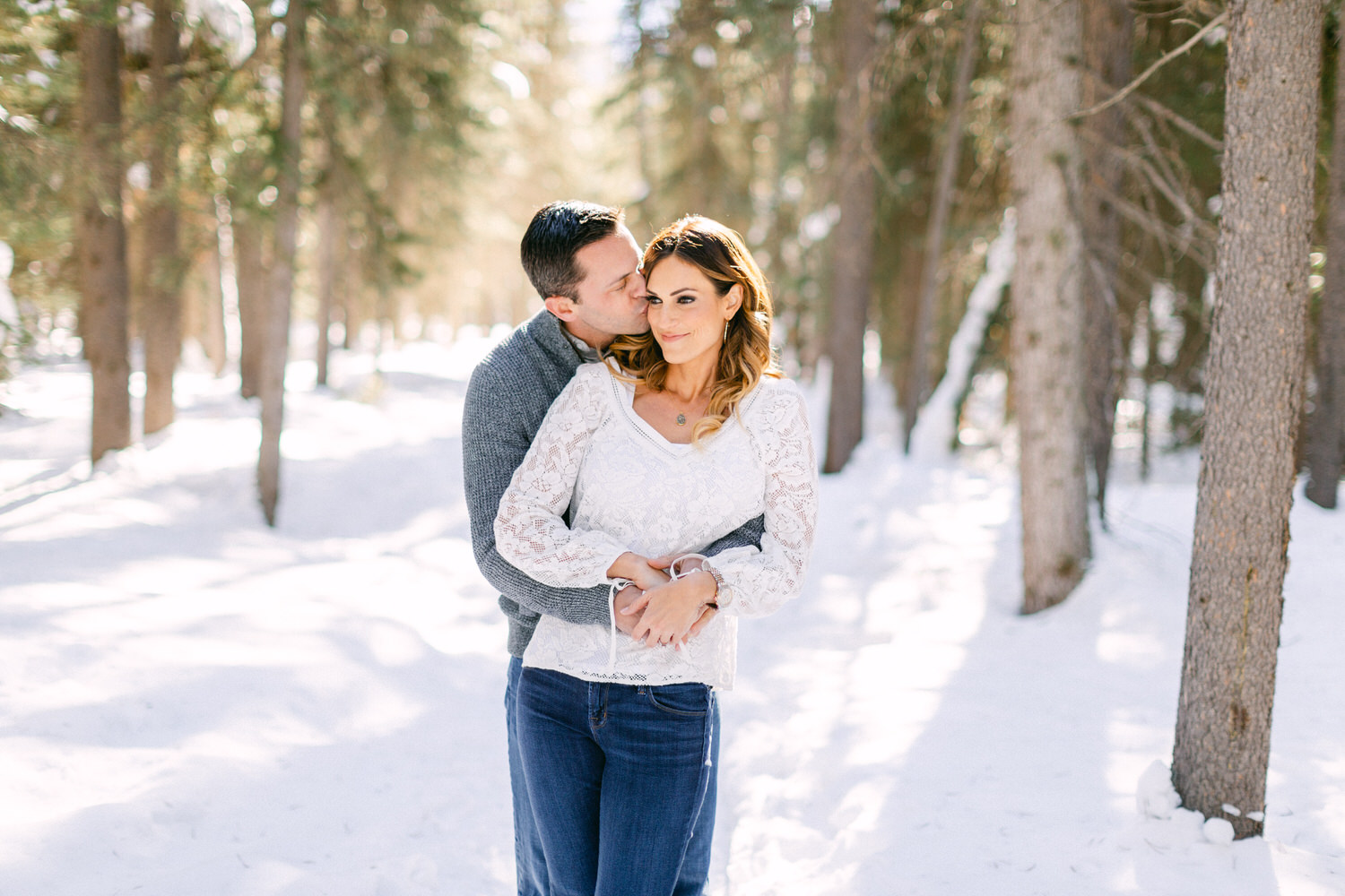 A couple hugging among snowy trees, the man kissing the woman's forehead while they both smile softly.