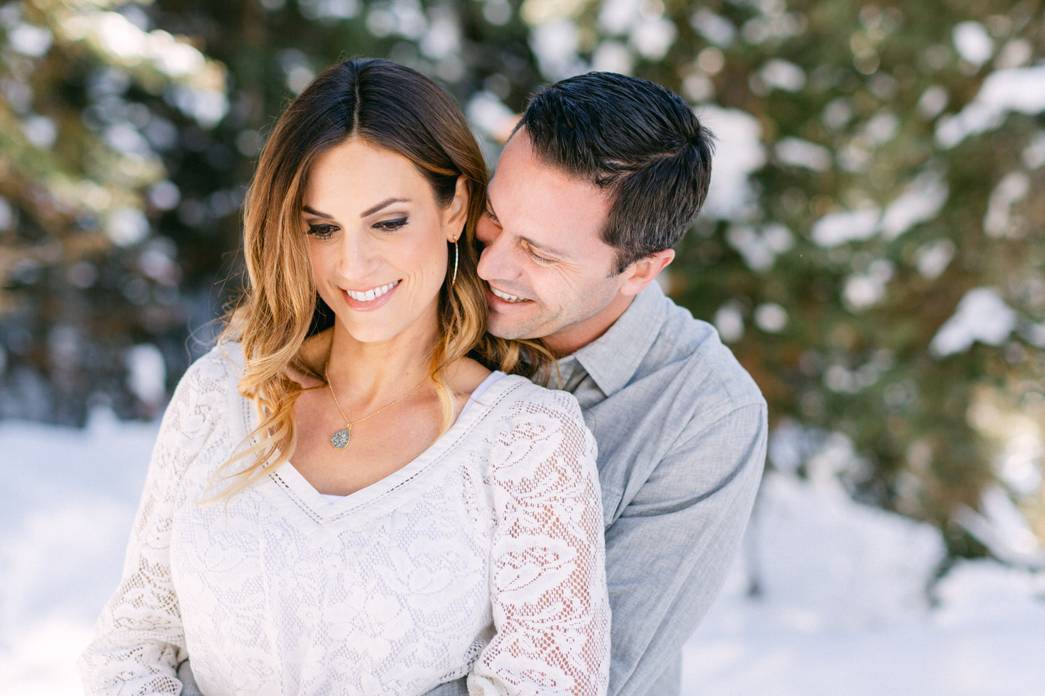 A smiling woman in a white sweater with a man hugging her from behind, both standing in a snowy setting with trees in the background.