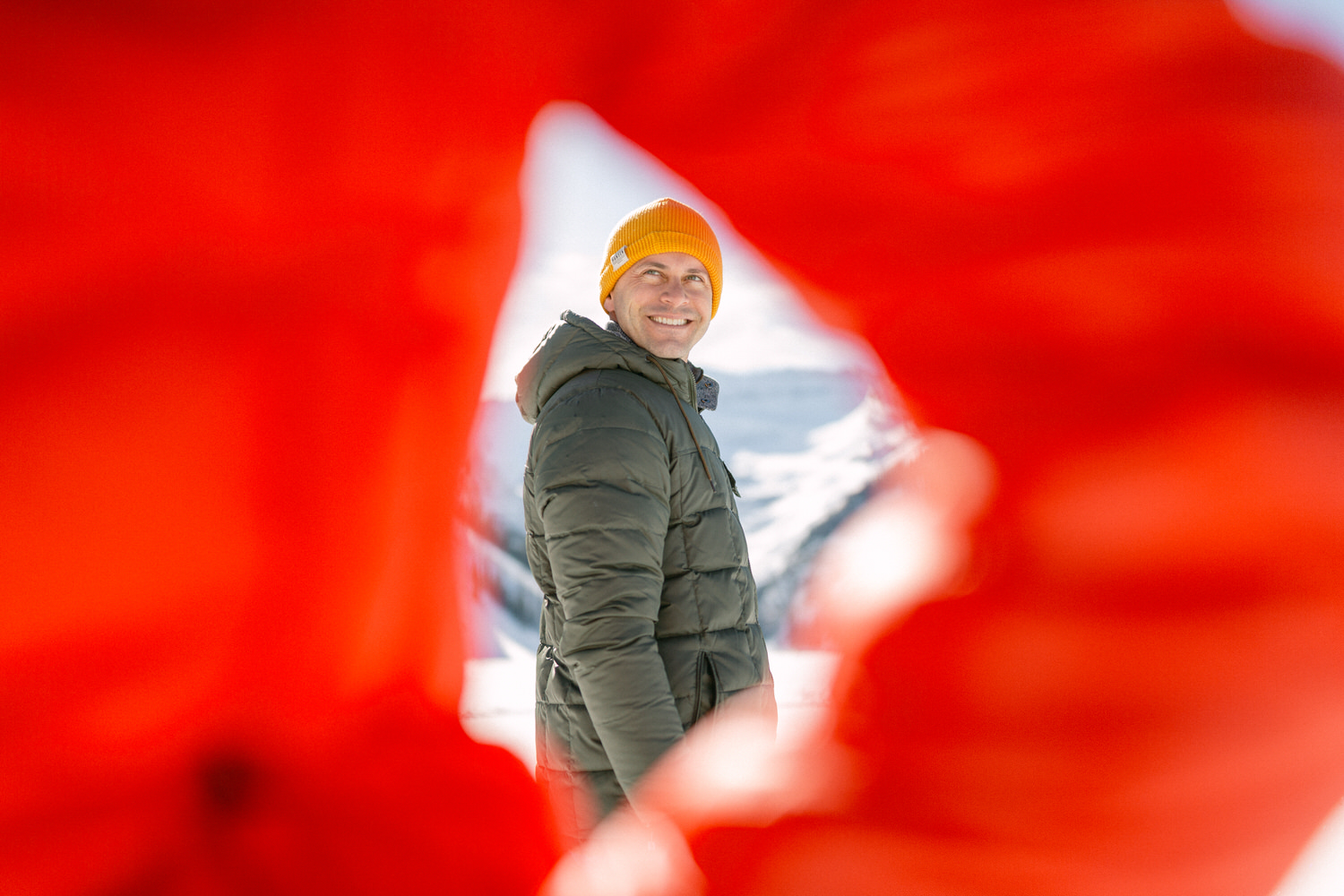 A cheerful man wearing a yellow beanie and a warm jacket seen through an out-of-focus red fabric foreground with snowy mountains in the background.