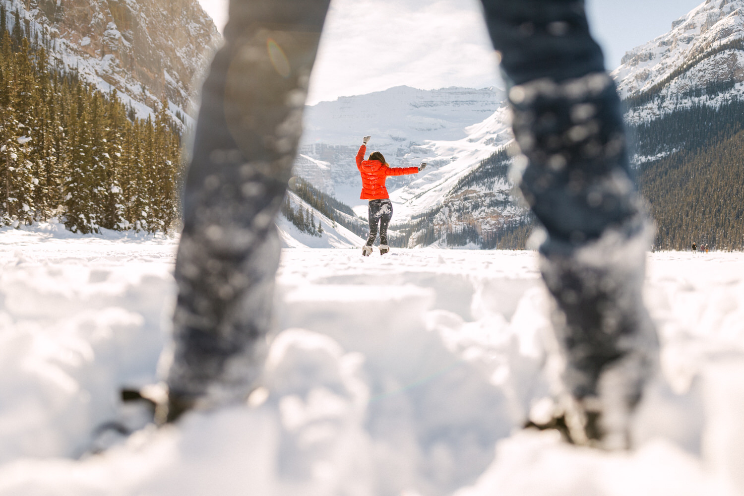 A person in a red jacket joyfully raises their arms amidst a snowy mountain landscape, viewed through a blurry foreground of snow and legs.