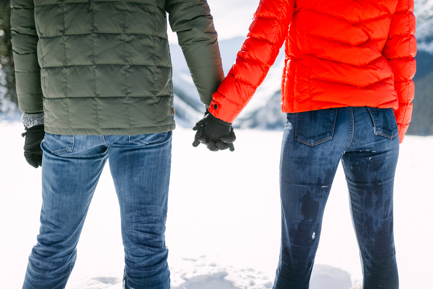 Two people holding hands wearing winter jackets and gloves in a snowy landscape