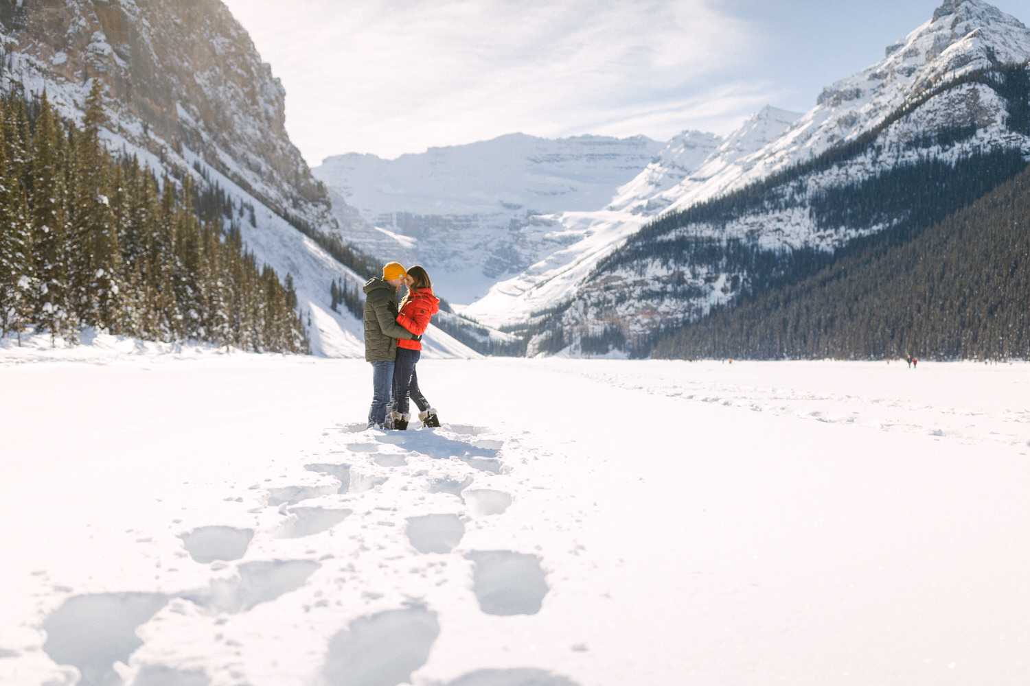 Two people embracing in a snowy landscape with mountains in the background and a trail of footsteps leading to them.