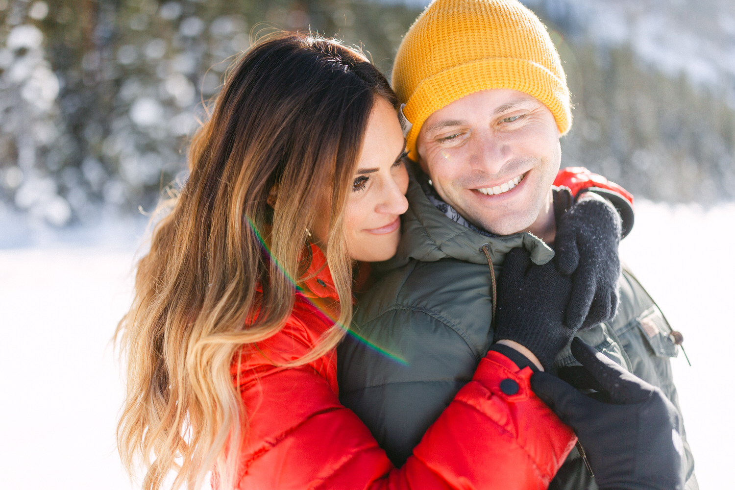 A smiling couple hugging in a snowy landscape, with the woman wearing a red jacket and the man in a green jacket and yellow beanie.