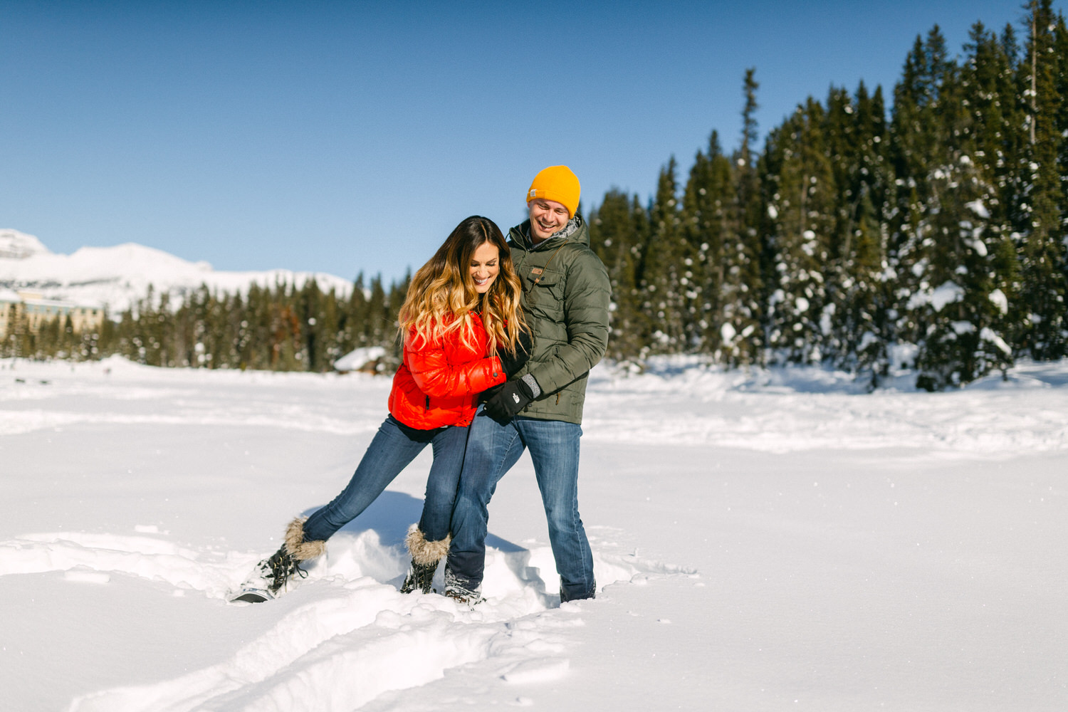 A couple playfully walking through deep snow with a forest and mountains in the background, sunny winter day.