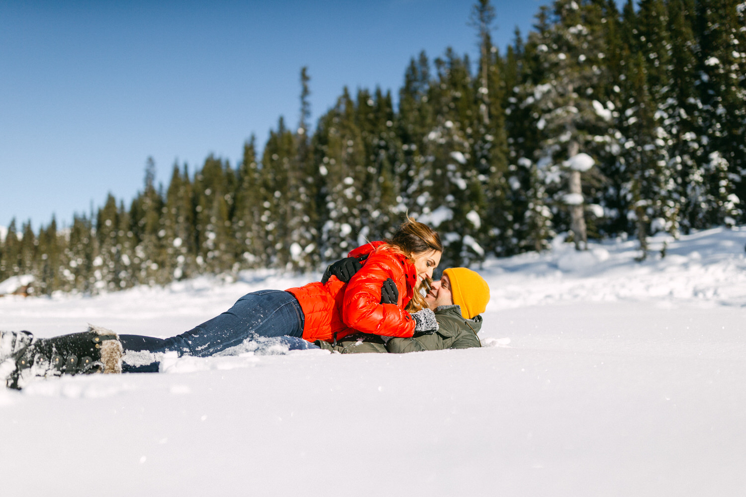 Two people sharing a tender moment lying in the snow, surrounded by a forest.