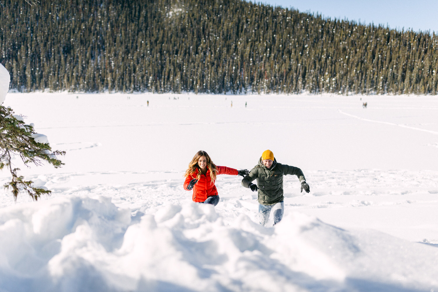 A man and a woman in winter clothing having a playful snowball fight on a snowy field with a forest in the background.