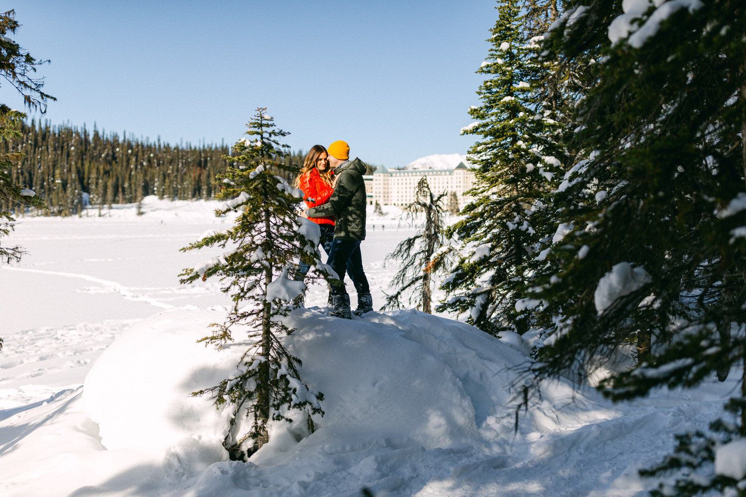 Two people standing among snow-covered trees with a clear sky and a building in the distant background.