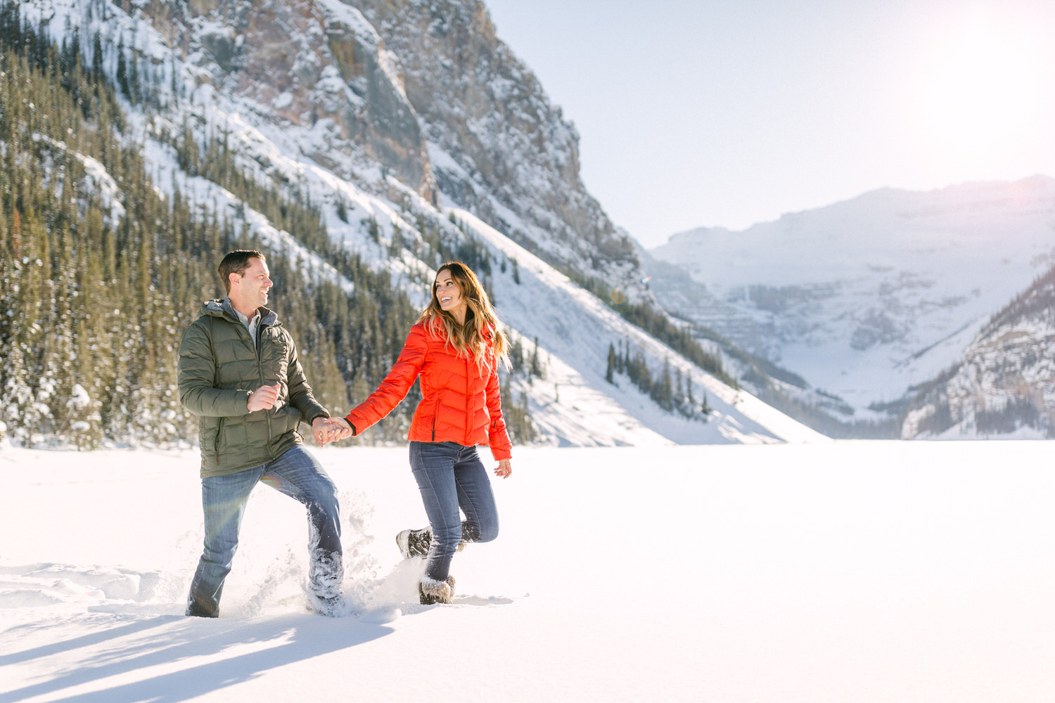 A couple holding hands and smiling, walking through a snow-covered landscape with mountains in the background.