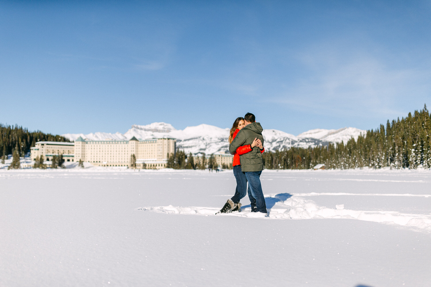 A couple hugging on a snowy field with a scenic mountain range and a large hotel in the background.