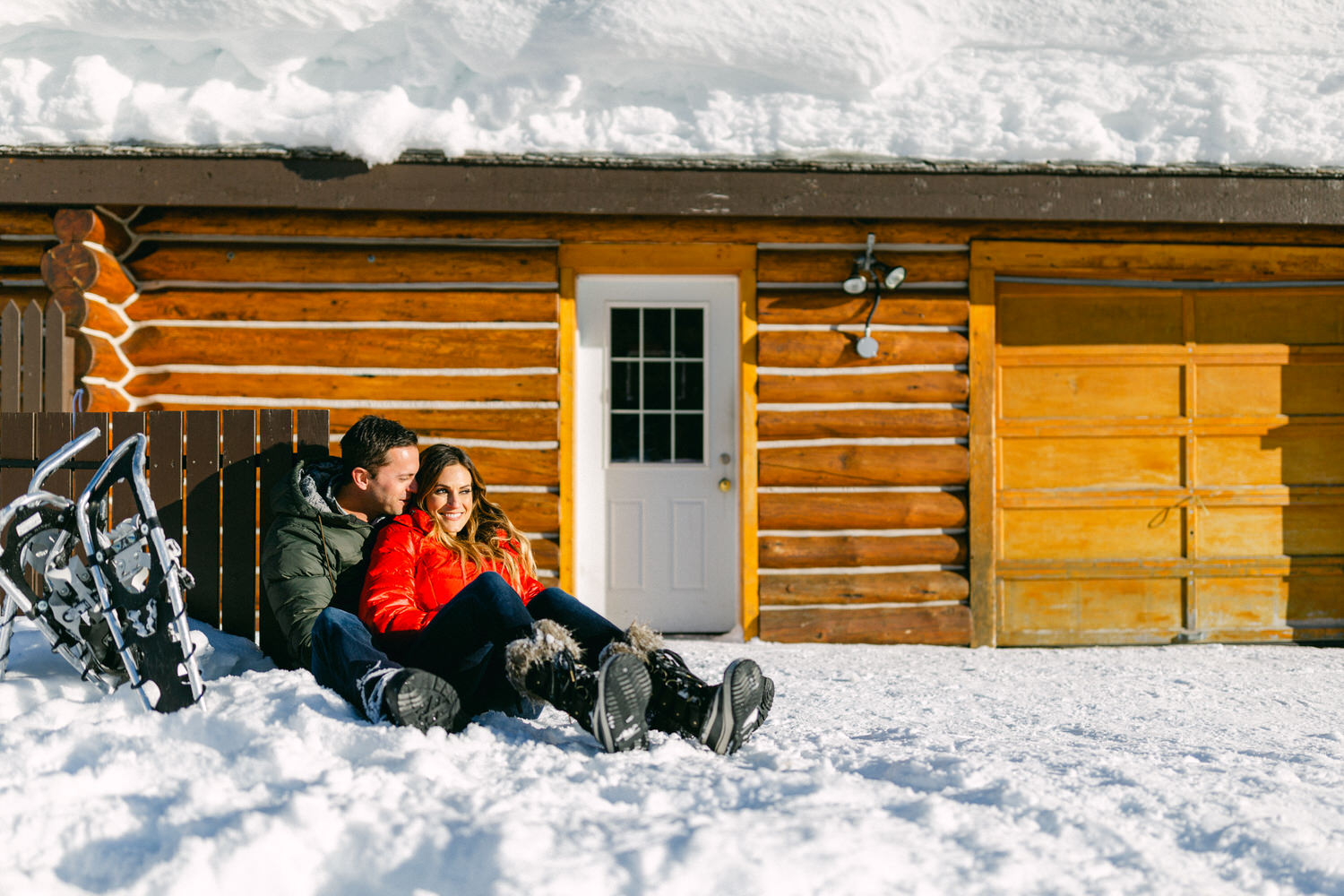 A couple sitting close together in the snow in front of a wooden cabin, with a pair of snowshoes nearby.