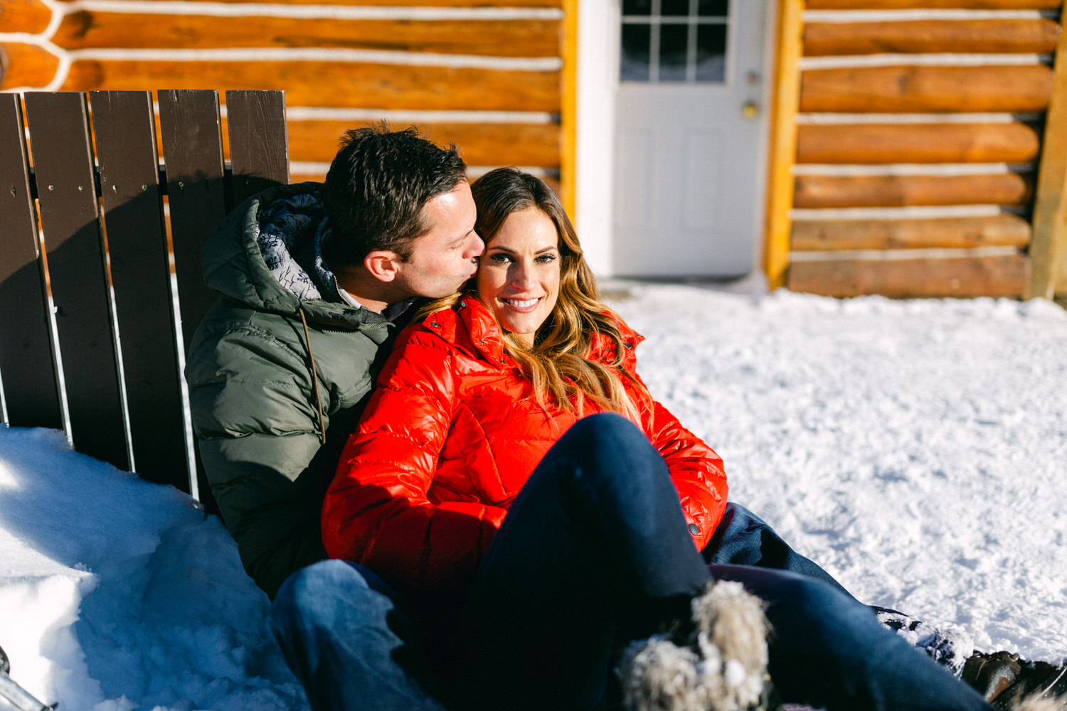 A couple sitting close together on a bench in the snow, with a man kissing the woman's cheek outside a log cabin.