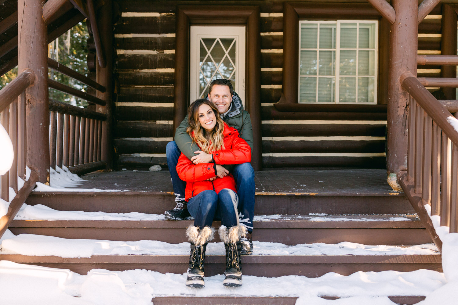 A smiling couple in winter attire hugging each other while sitting on snow-covered steps of a wooden cabin.