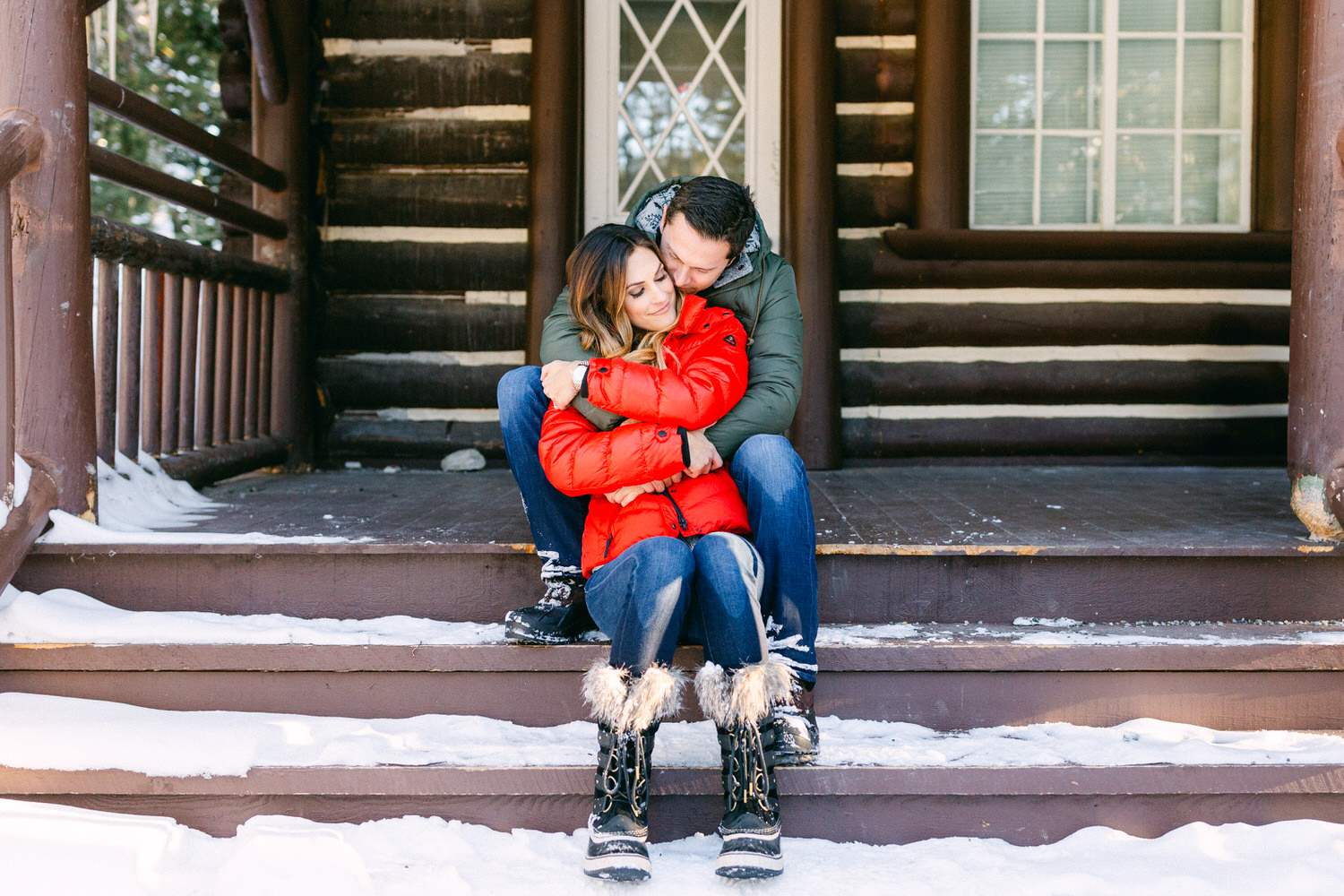 A couple hugging each other while sitting on the steps of a snow-covered porch of a cabin.