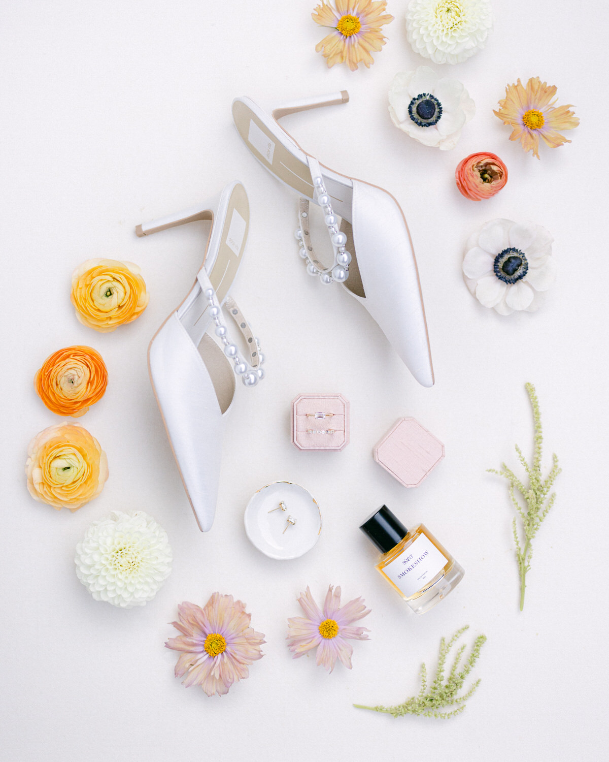 A pair of white bridal shoes adorned with pearls, surrounded by colorful flowers, a ring on a small dish, a jewelry box, and a bottle of perfume arranged on a pale background.