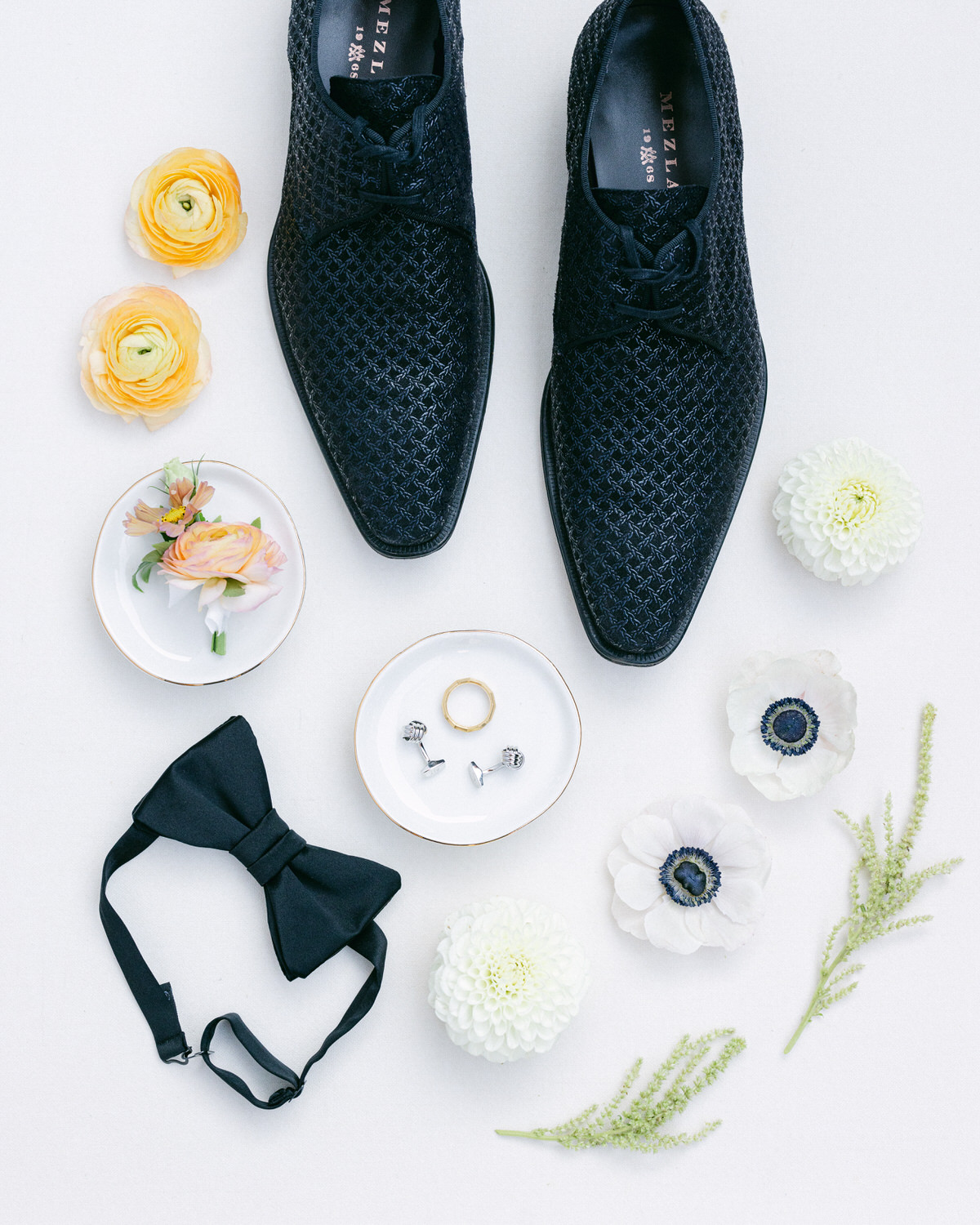 A pair of textured black formal shoes, a bow tie, cufflinks, a wedding ring, and a selection of flowers and greenery artfully arranged on a light background.