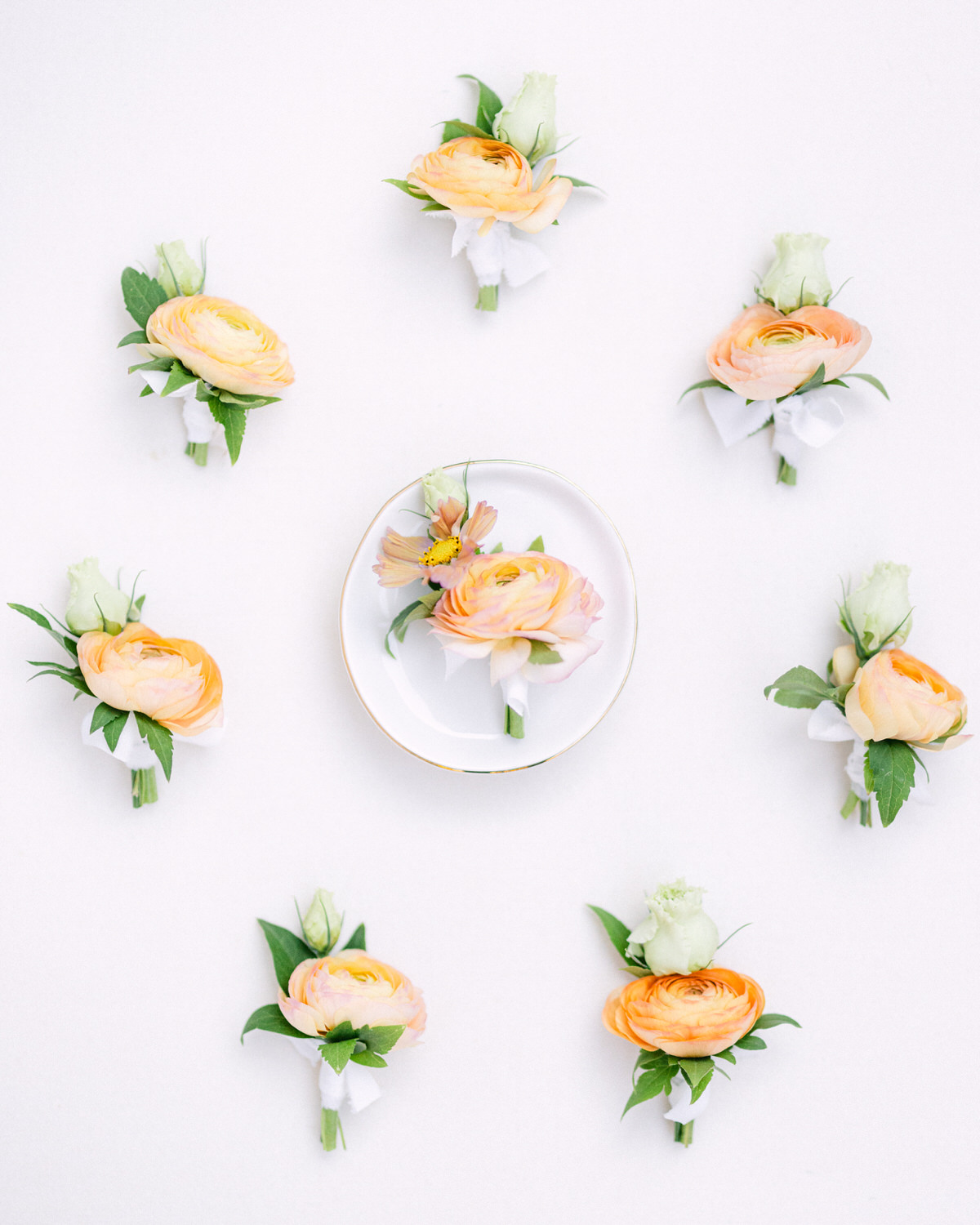 An overhead view of a symmetrical arrangement of peach ranunculus flowers and boutonnieres on a light background, with a plate adorned with a flower in the center.