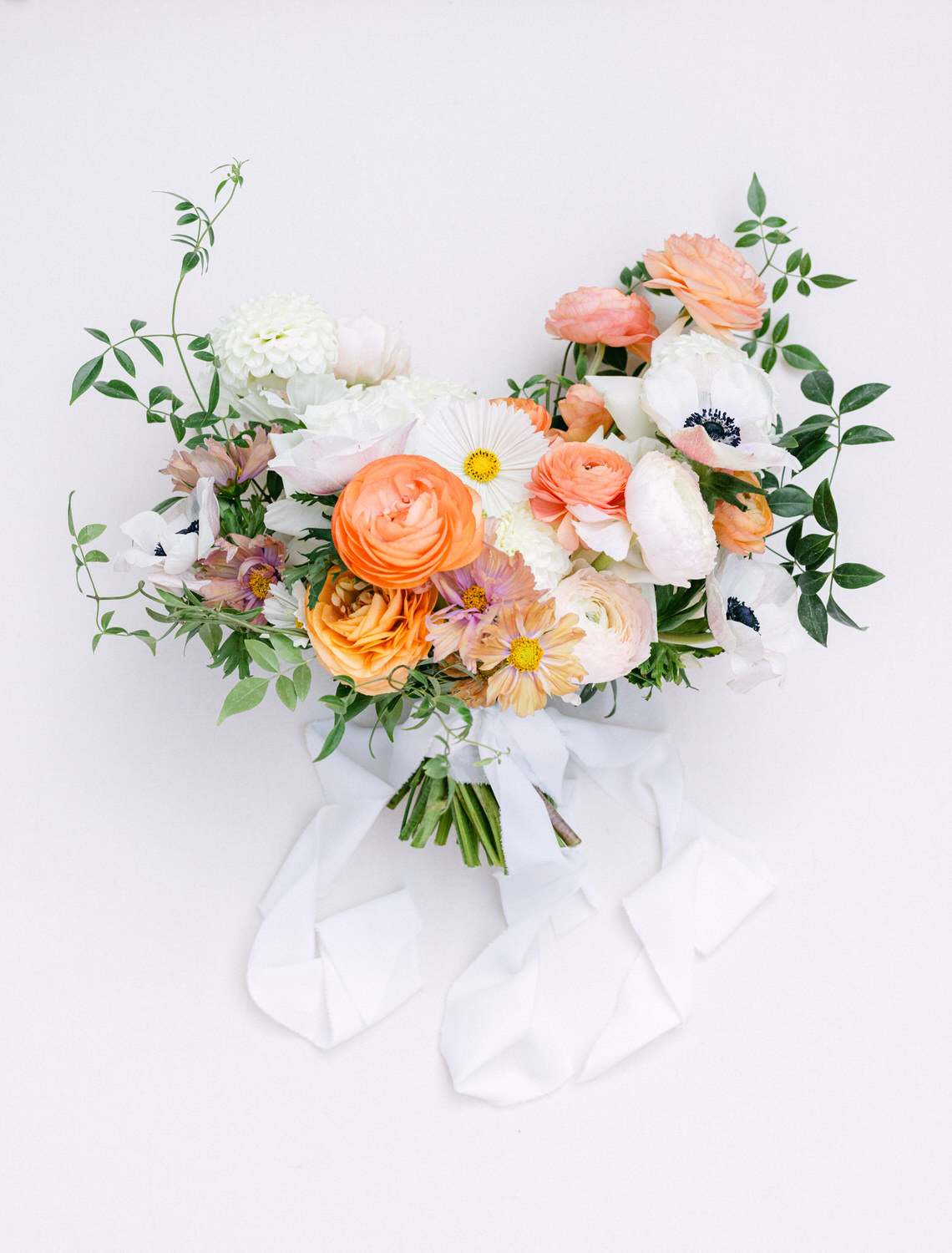 A beautifully arranged wedding bouquet with orange, white, and pink flowers, including roses and daisies, accented by green foliage and long white ribbons against a light background.