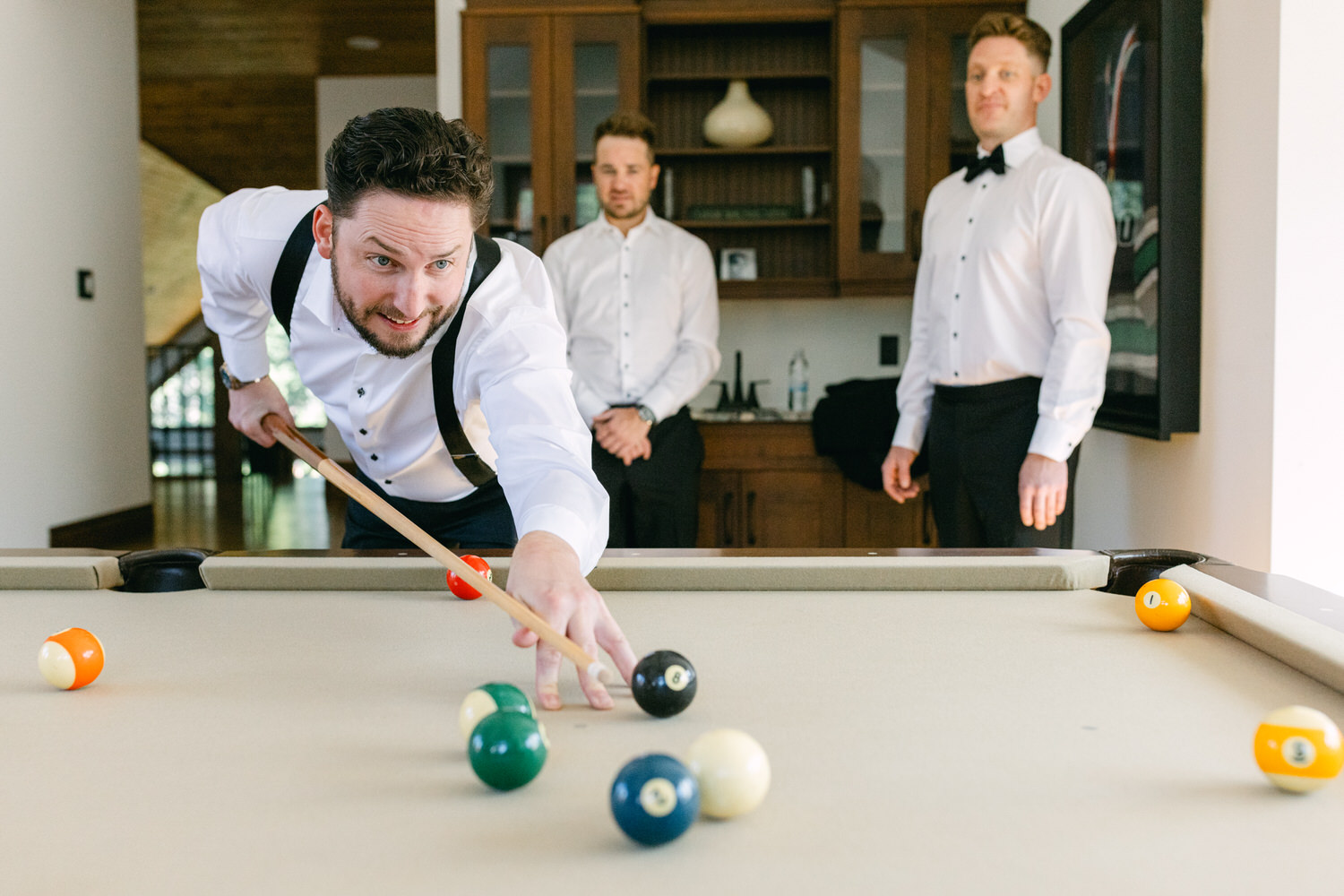 A man in a vest and dress shirt aiming on a billiard table while two others watch.