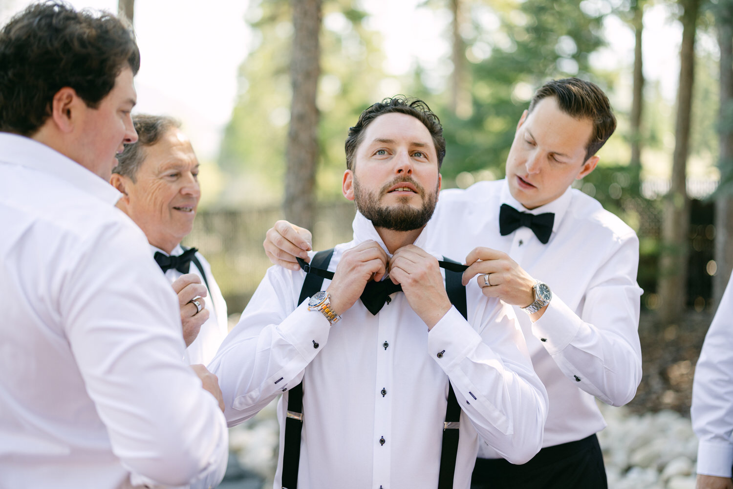 A group of men wearing white shirts with suspenders and black bow ties, adjusting the groom's attire in a forest setting.