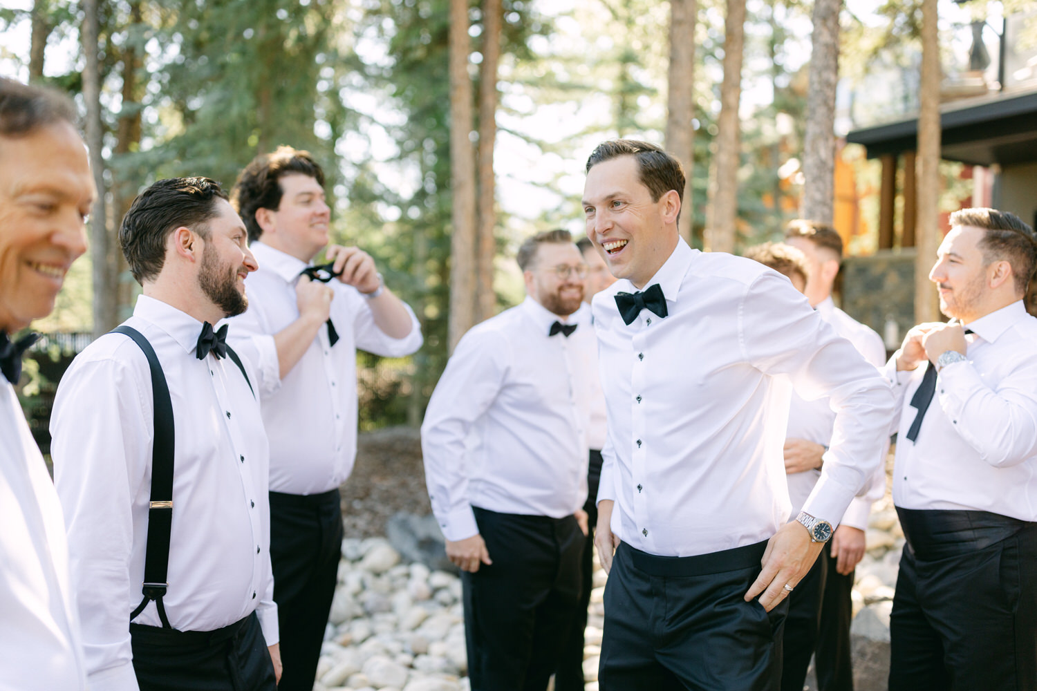 A group of men in formal attire with bow ties and suspenders, smiling and chatting in an outdoor setting.