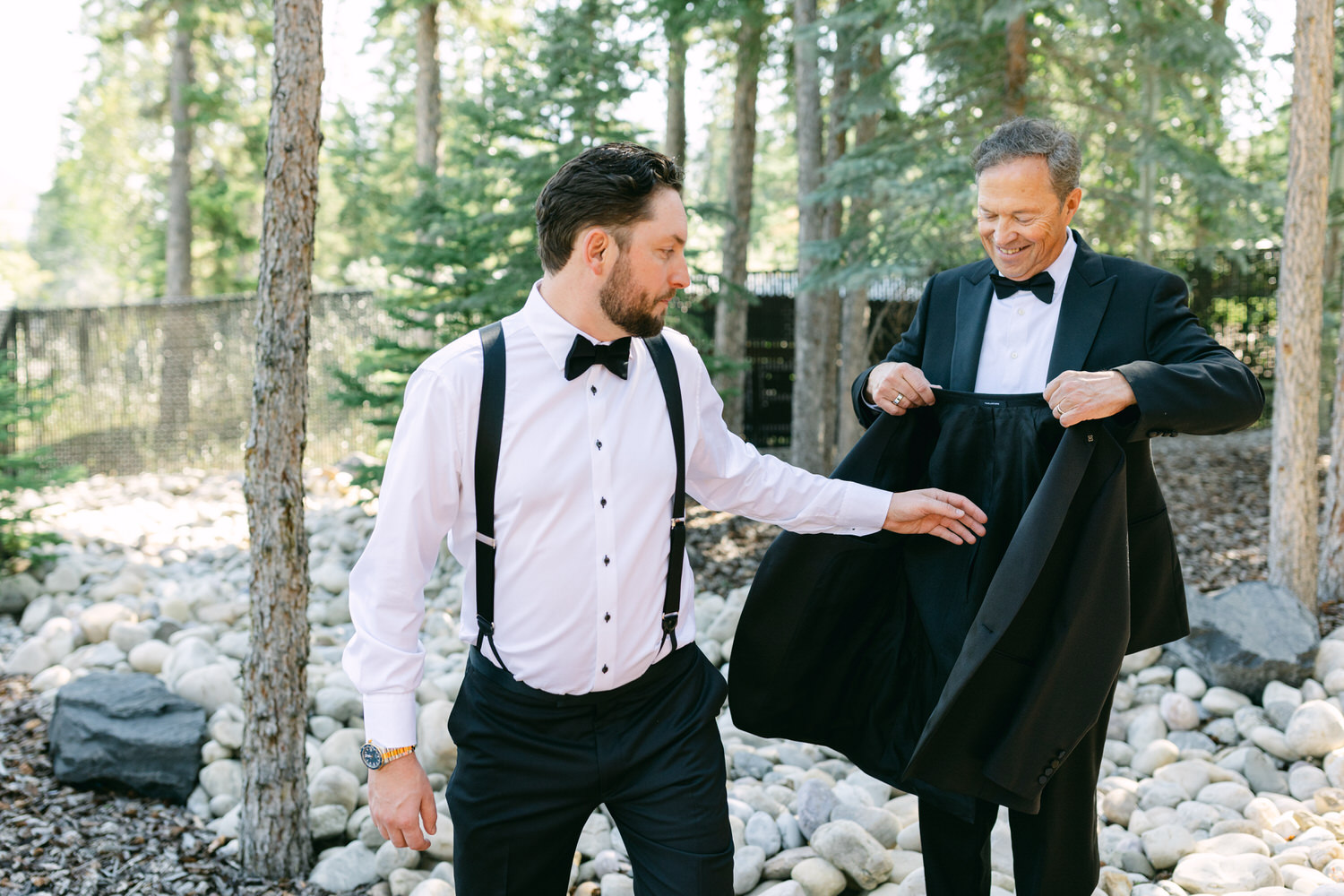 Two men in formal attire with one adjusting the other's tuxedo in a forest setting.